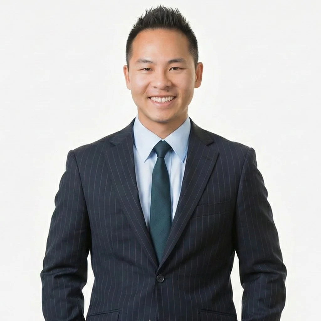 A young Asian man in a dark pinstripe suit, light blue shirt, and dark tie, smiling and standing against a plain white background.