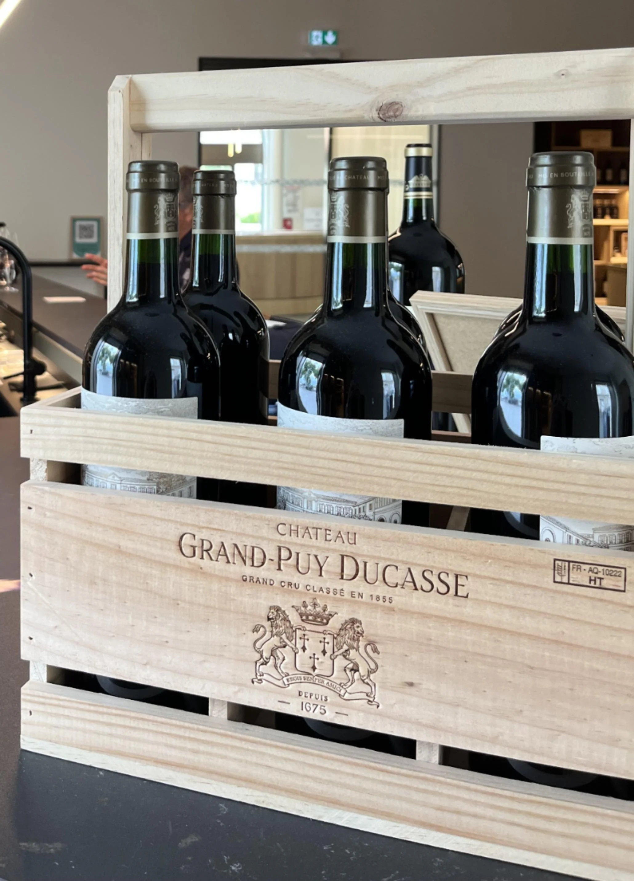 Wooden crate labeled 'Chateau Grand-Puy Ducasse' containing bottles of wine, with glossy dark glass bottles and gray metal caps.