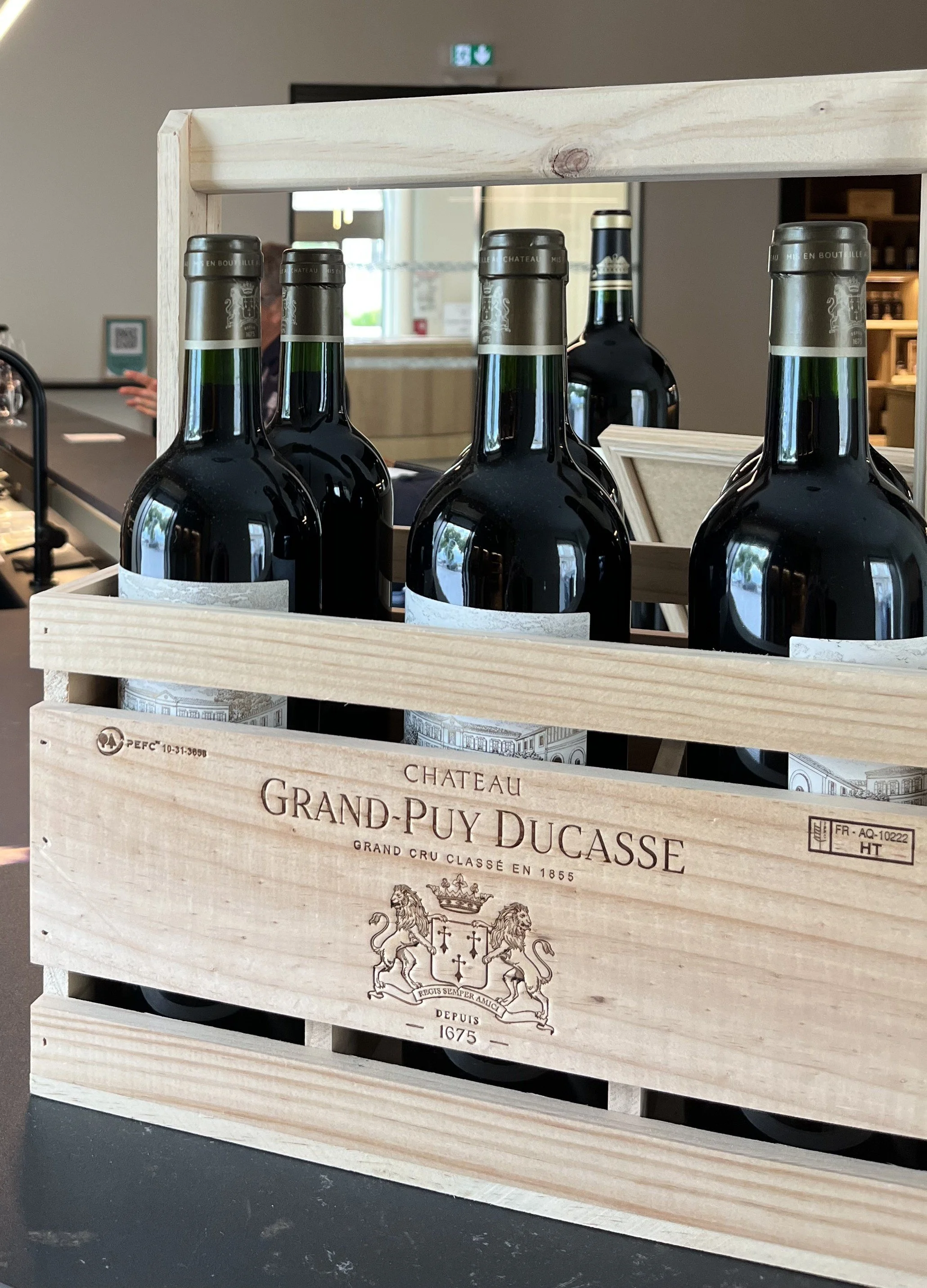 A wooden crate labeled Chateau Grand-Puy Ducasse holding several bottles of red wine at a store or cellar.