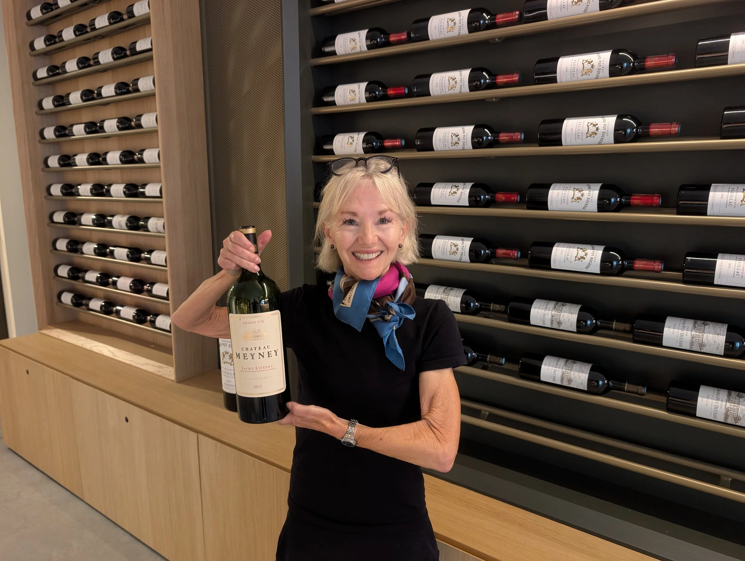 Smiling woman with blonde hair holding a bottle of red wine in front of wine racks filled with bottles of wine.