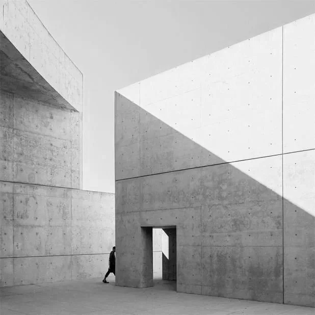 A person walking through an architectural concrete structure with minimalist design and geometric shapes, featuring a large doorway and distinct shadows.