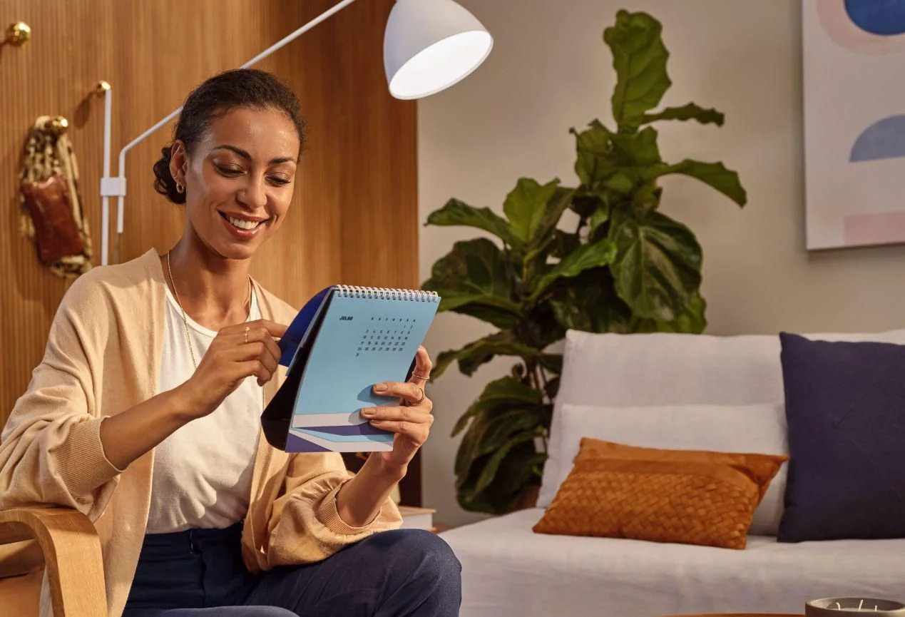 Mulher sorridente sentado na sala, usando roupas casuais, interagindo com um telefone ou dispositivo eletrônico, com uma planta grande ao fundo e uma parede decorada