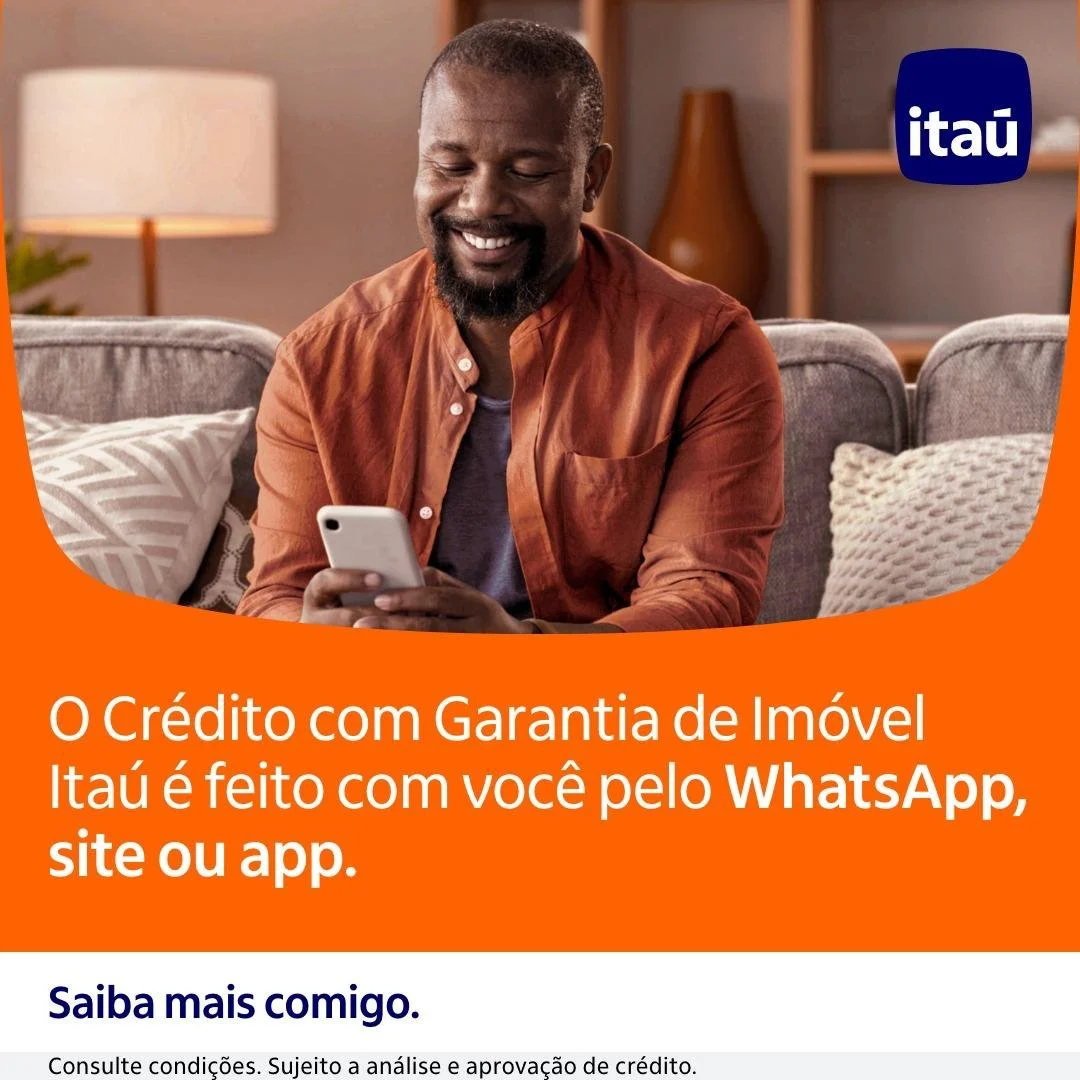 Homem sorridente usando celular em sala de estar moderna com móveis e luminária de parede, promovendo crédito com garantia de imóvel do Itaú através de Whatsapp, site ou app.