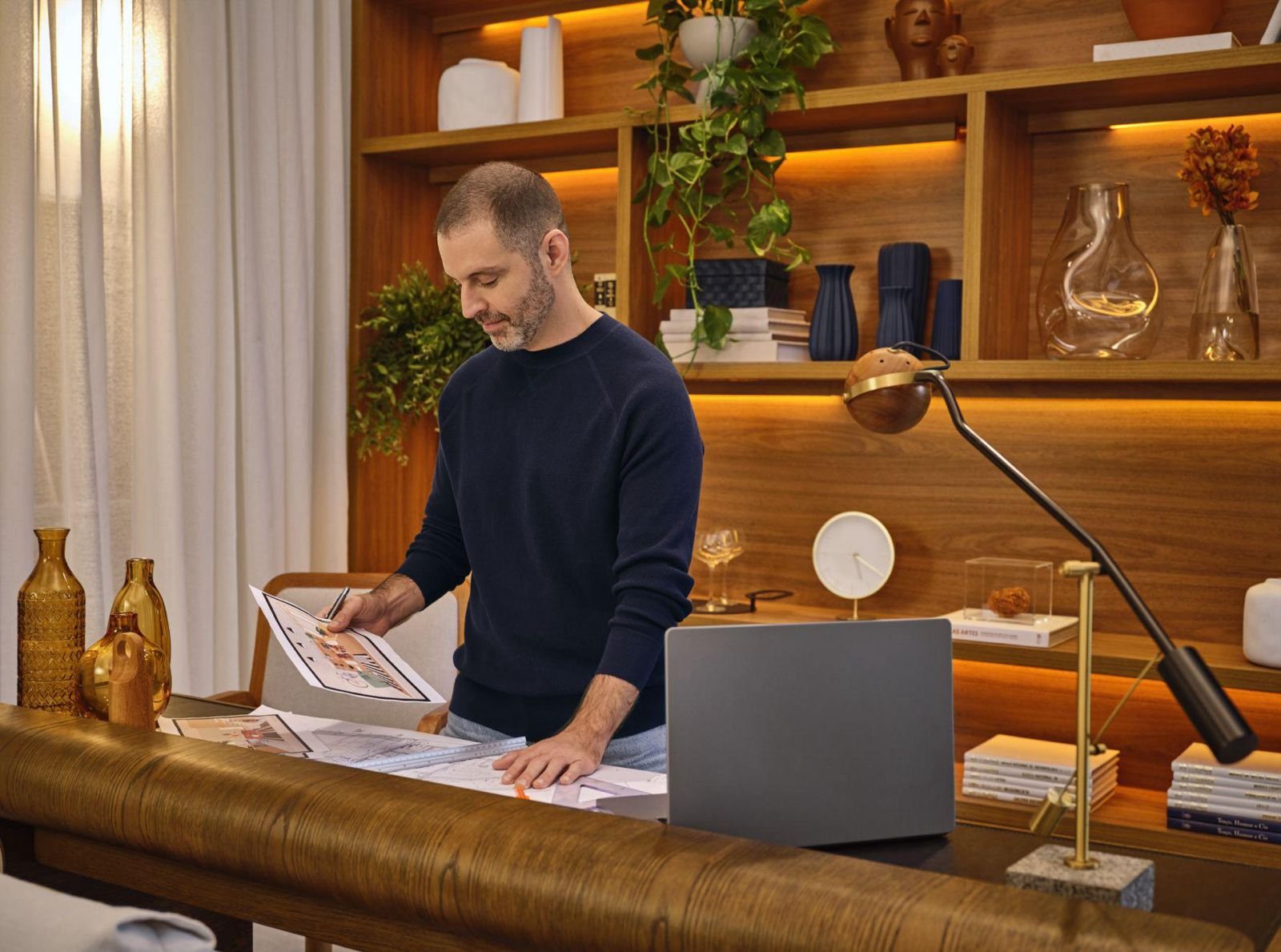 Homem em um escritório moderno, analisando papéis e projetos arquitetônicos em uma mesa de madeira, com livros e objetos de decoração ao redor, parede de madeira com prateleiras e plantas ao fundo.