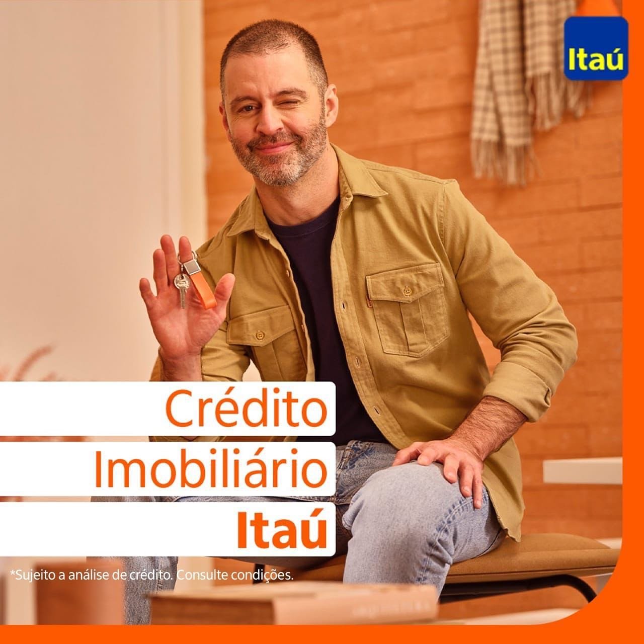 Homem sorridente segurando uma chave, promovendo crédito imobiliário do Itaú, com fundo de parede de madeira.