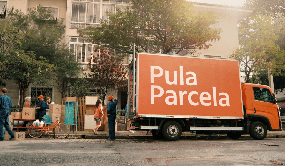 Caminhão de mudança amarelo com placa em português "Pula Parcela" na rua, com pessoas, caixas de papelão, bicicleta, árvores e prédio ao fundo