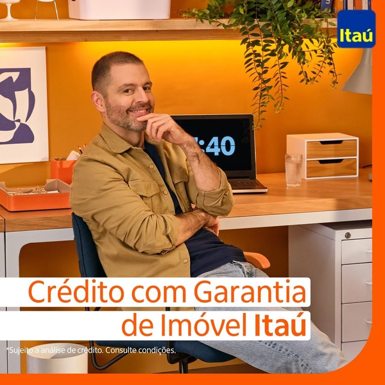 Homem sentado em uma mesa de escritório, sorrindo, com uma parede laranja ao fundo, um laptop, uma planta, uma gaveta e uma garrafa de água ao lado. O anúncio é do Itaú, promovendo crédito com garantia de imóvel.