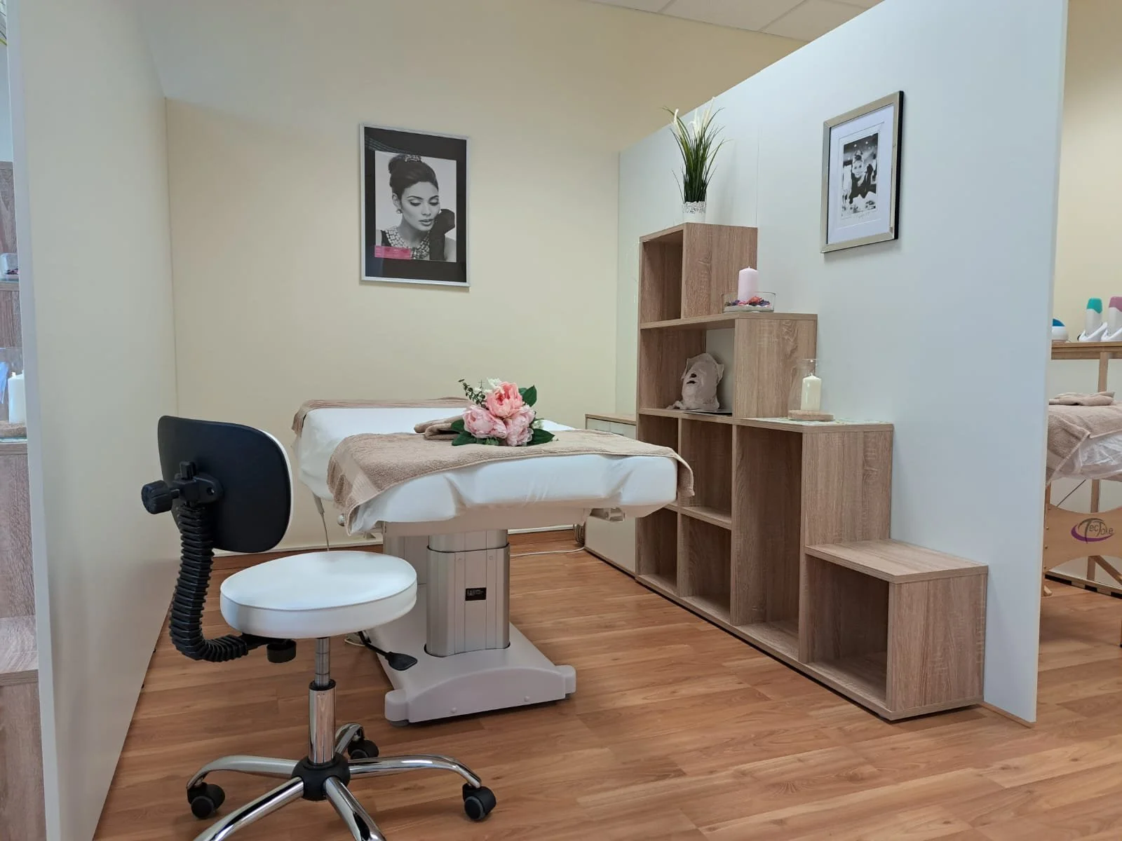 Ein Behandlungstisch in einem Wellness- oder Kosmetikstudio, dekoriert mit einer Blumenstrauß und einer beige Decke. Im Hintergrund sind Bilder an der Wand und ein Regal mit Dekorationsartikeln und Pflanzen.