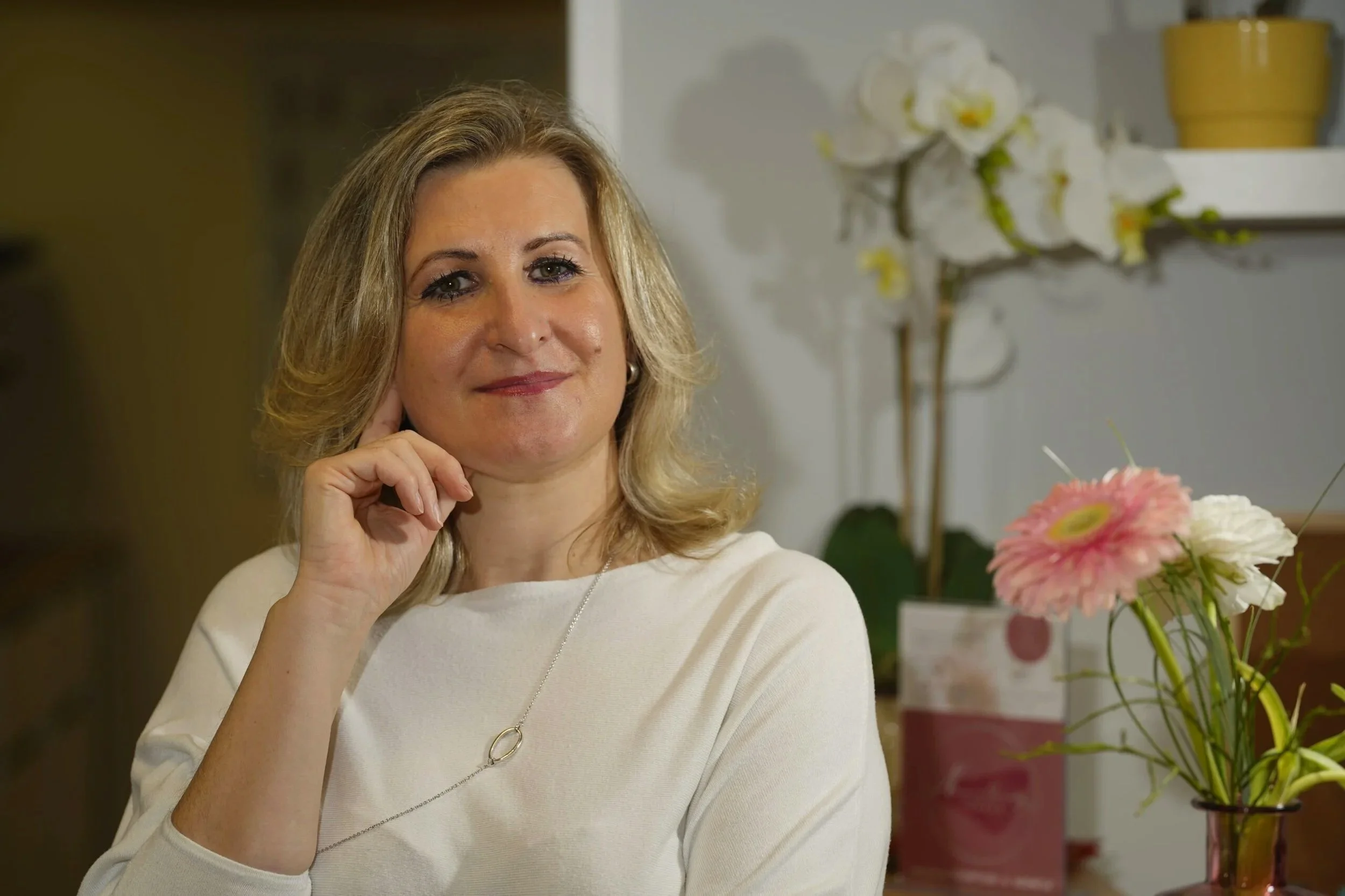 Eine lächelnde Frau mit blonden Haaren in einem hellen Pullover sitzt neben einem Blumenstrauß und einer Orchidee auf einer weißen Ablage.