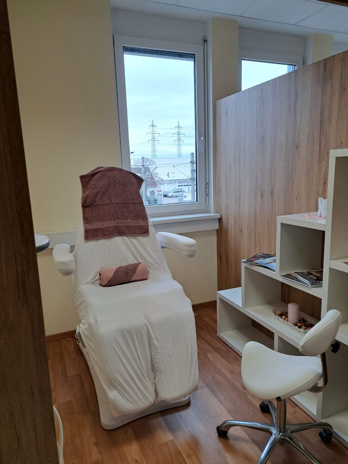Ein Behandlungsraum mit einem verstellbaren Stuhl, einem weißen Regal mit Zeitschriften und Dekorationsartikeln, und einem Fenster mit Blick auf Stromleitungen und die Außenwelt.