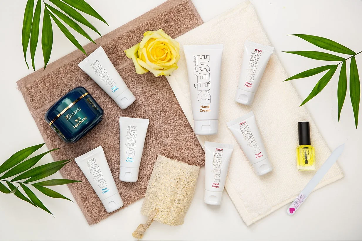 Pflegeprodukte auf Handtüchern mit grünen Blättern, darunter Lotionen, Creme, Nagellack, Nagelknipser, Schwamm und Rose.