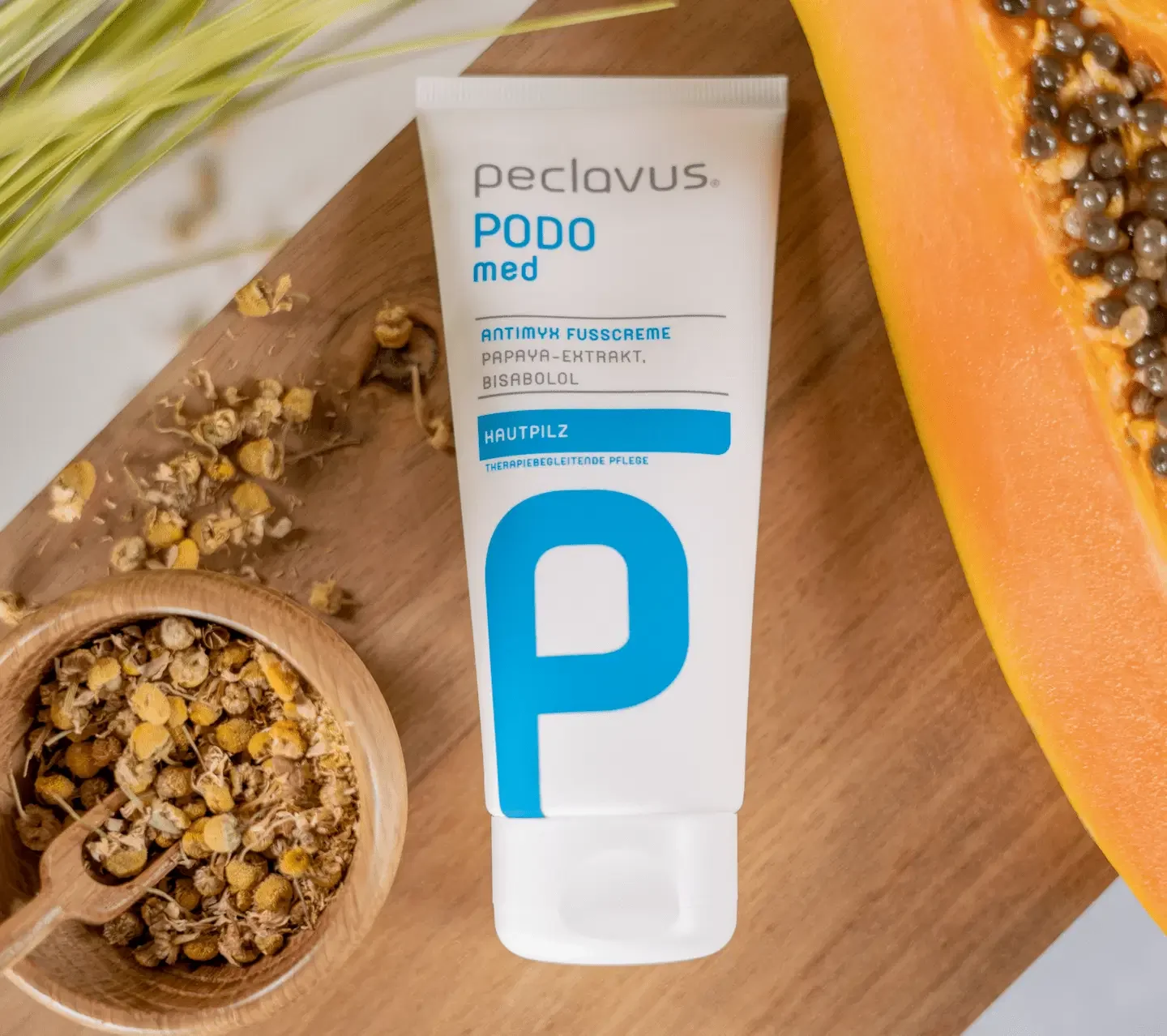 Tube mit Peclavus Podo med antimykotischer Fußcreme neben einer Papaya und Kamille auf einem Holztisch.