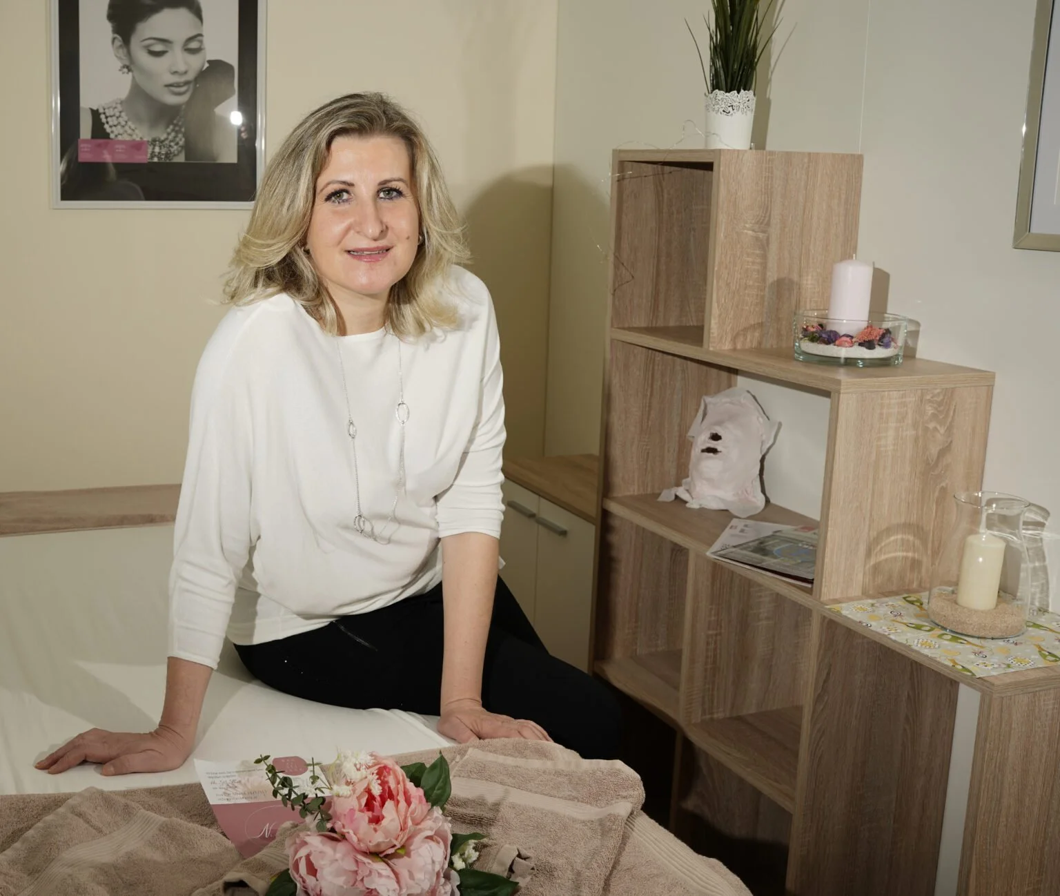 Eine Frau sitzt auf einem Bett in einem Raum mit Holzmöbeln. Im Hintergrund befindet sich eine schwarze und weiße Wanddekoration. Auf dem Bett liegen Blumen und ein Grußkarton.