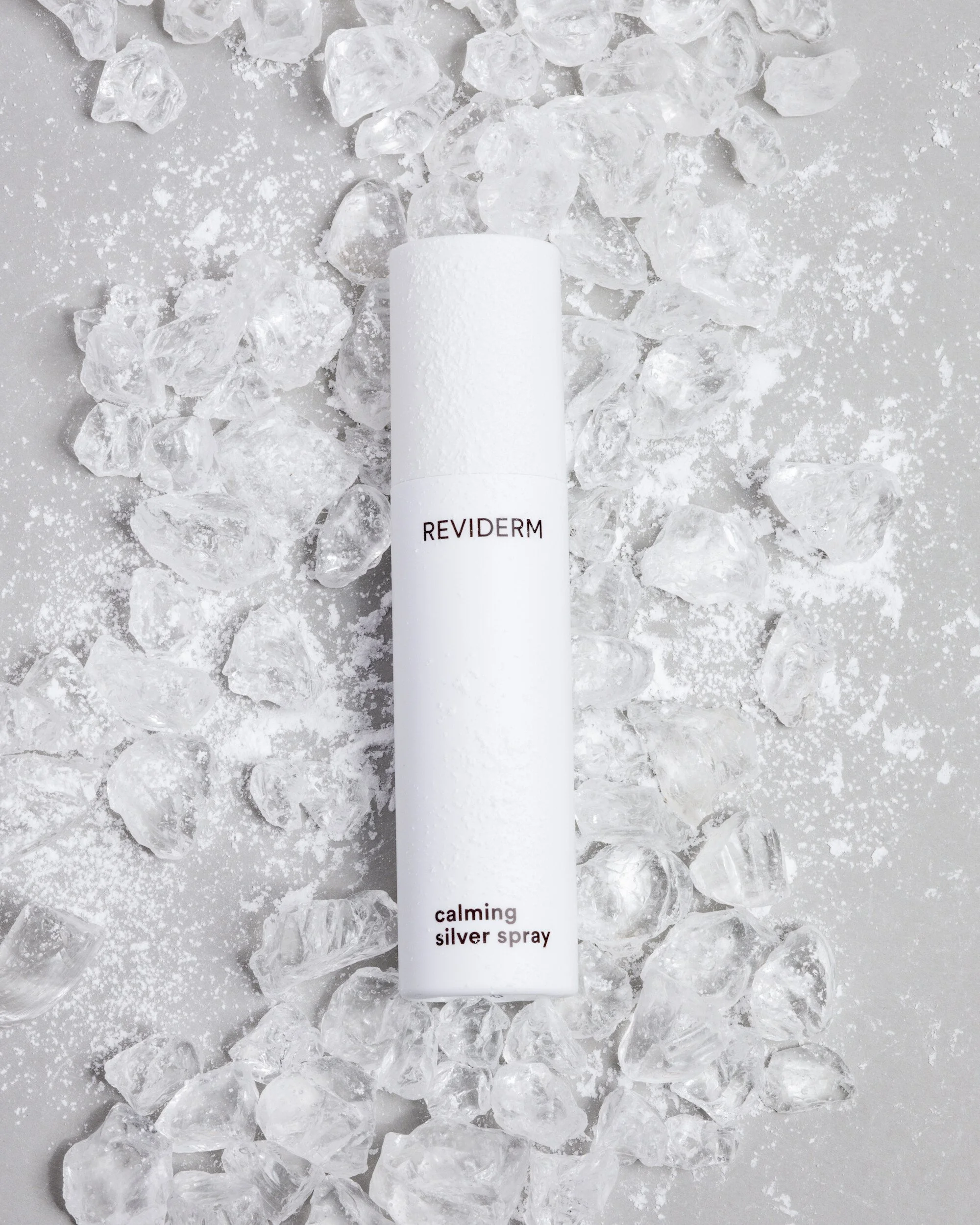 Ein weißer Spray mit der Aufschrift 'REVIDERM calming silver spray' liegt auf einer Oberfläche mit verstreuten Eiswürfeln und Puderzucker.