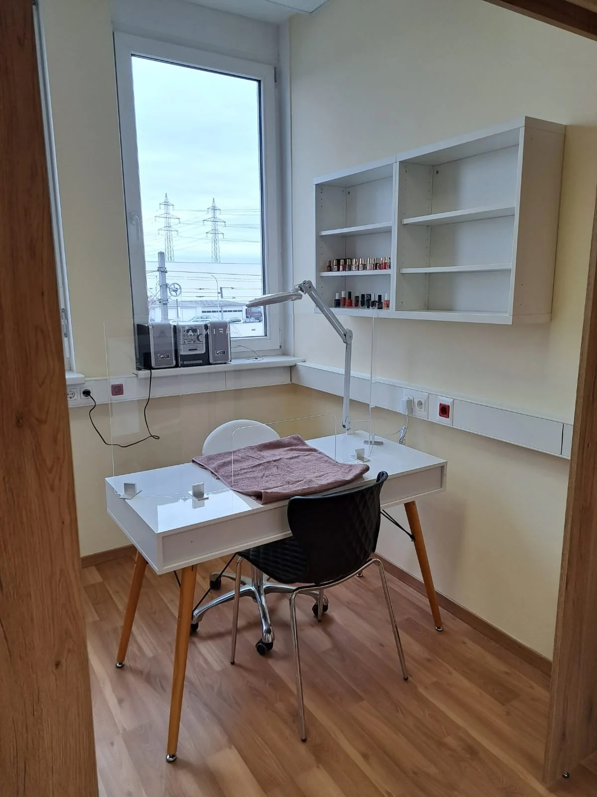 Ein Arbeitsplatz mit einem weißen Tisch, einem schwarzen Stuhl, einer Lampe, einem Handtuch und Nägel auf dem Tisch. Es gibt eine Fenster mit Sicht auf Strommasten und ein Radio auf der Fensterbank. An der Wand ist eine weiße offene Regal mit Nagella