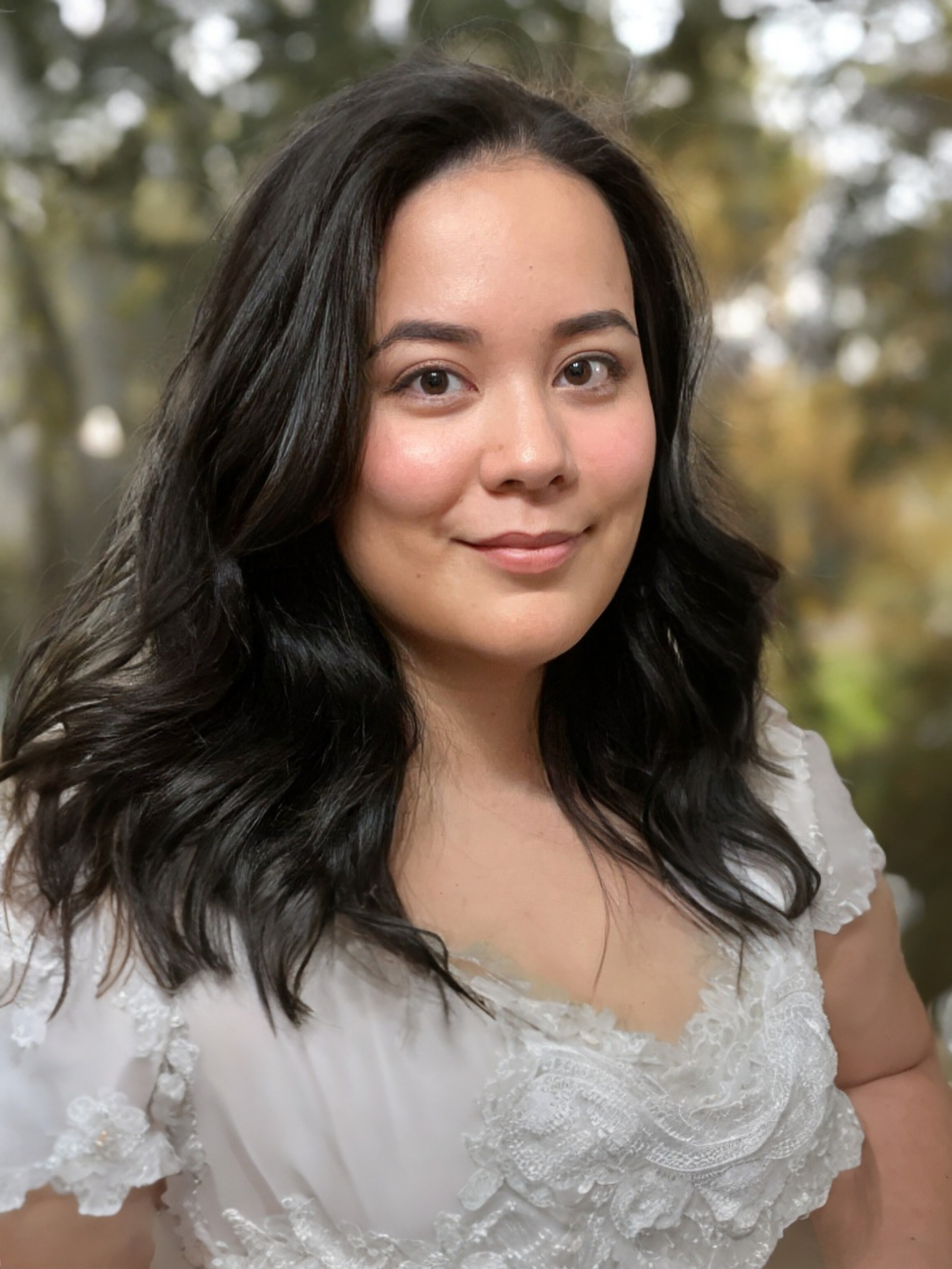 A young woman with long dark hair, smiling softly on an outdoor background with blurred trees.