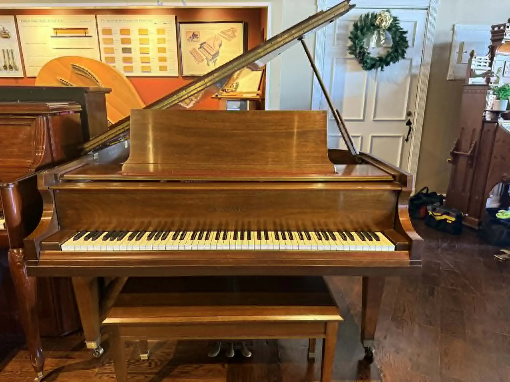A wooden upright piano with its lid open, displaying the strings inside, in a room decorated with a Christmas wreath on the door and musical charts on the wall.