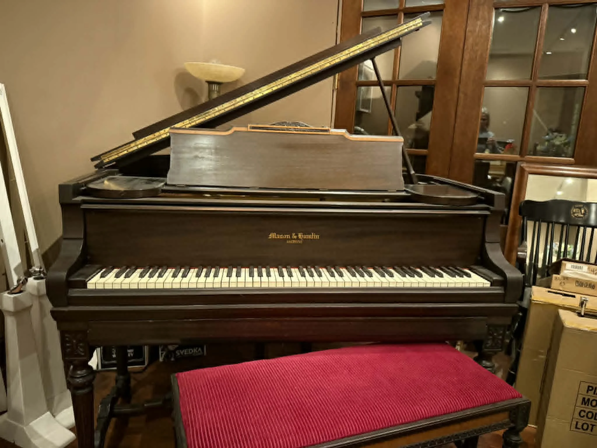 A vintage brown Steinway & Sons upright piano with open lid, showing strings and hammers, situated in a room with wooden furniture and a red upholstered bench in front.