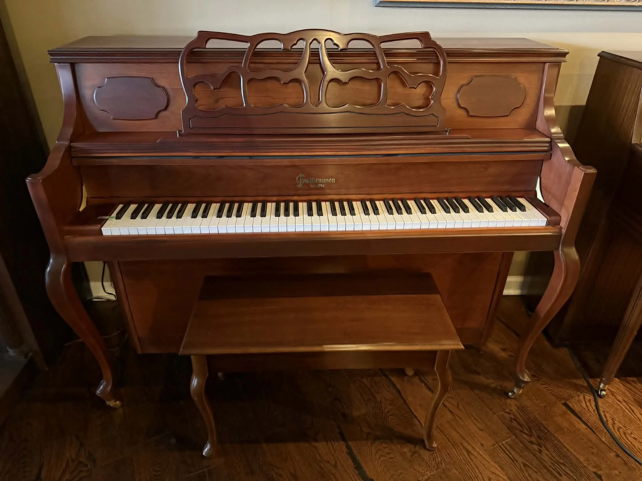 A vintage wooden upright piano with a matching bench, ornate music stand, and a brand name 'Gulbransen' on the front, in a room with hardwood floors.