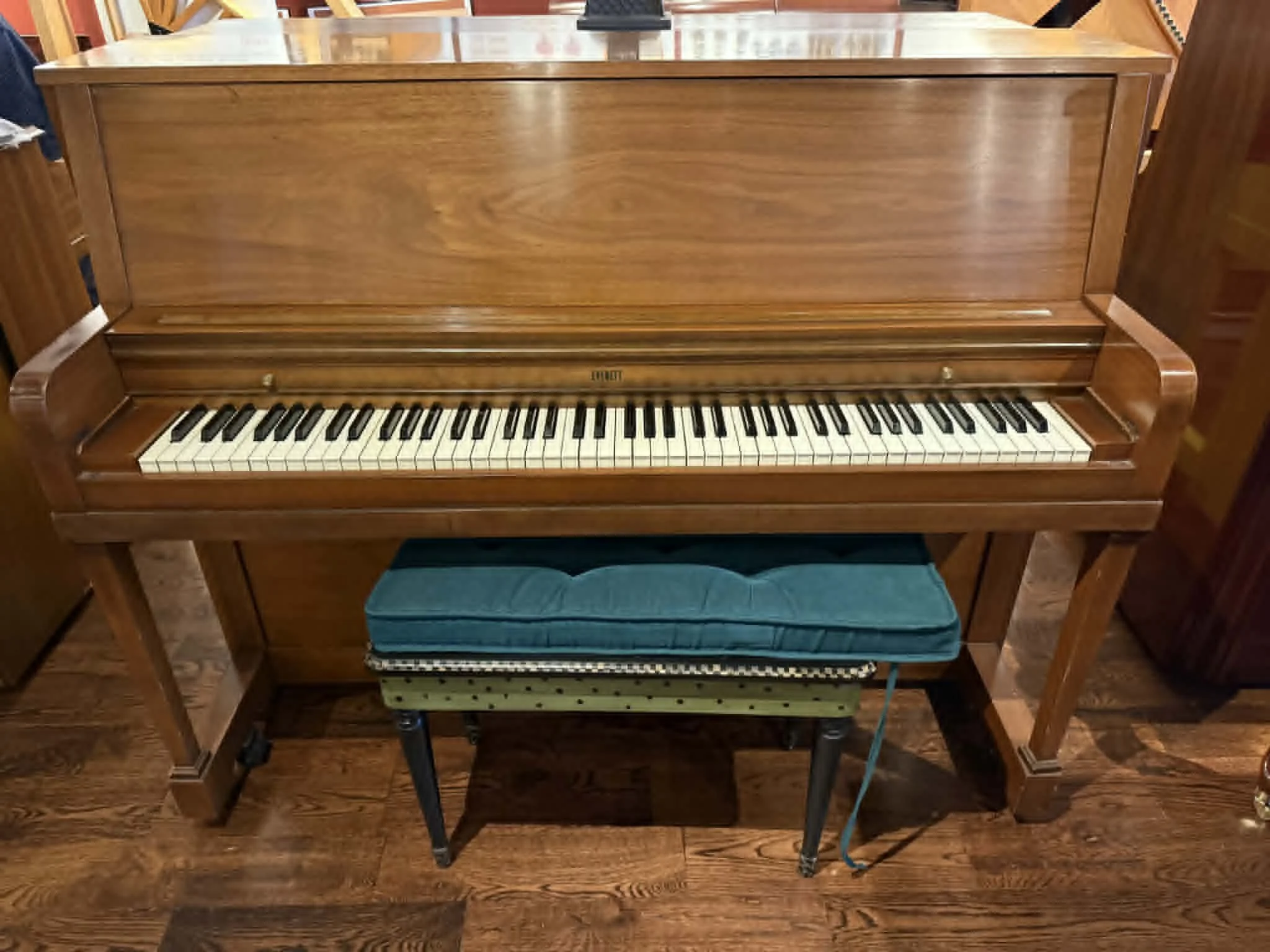 A vintage upright brown wooden piano with a bench featuring teal padding and black legs, situated on a hardwood floor.