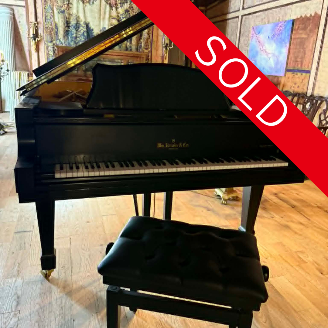 A black grand piano with the lid open, placed on a wooden floor in a room with wooden walls and colorful art on the walls. A bench is in front of the piano. The image has a red diagonal banner with white text that says 'SOLD.'