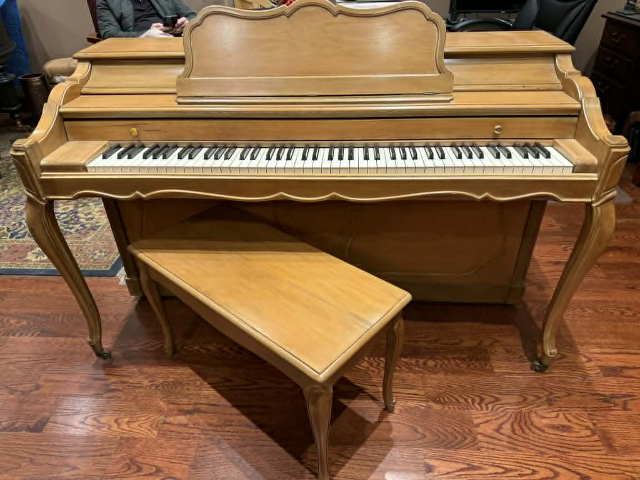 A vintage wooden upright piano with a matching bench, situated on a hardwood floor in a room with people and furniture in the background.