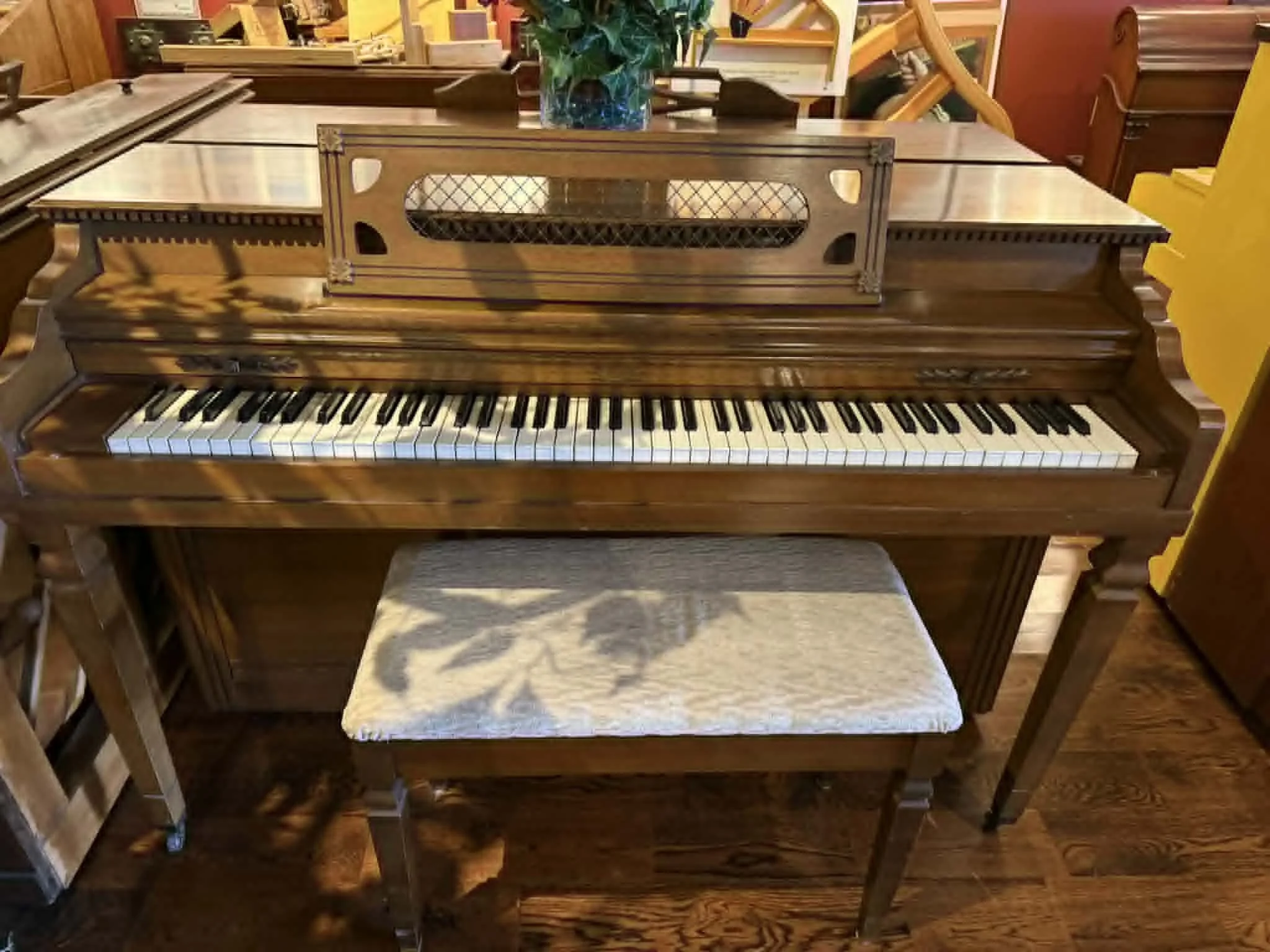 A vintage upright piano made of dark polished wood with a sheet music stand on top, positioned in a room with wooden flooring and furniture. There is a cushioned bench with a patterned fabric in front of the piano.