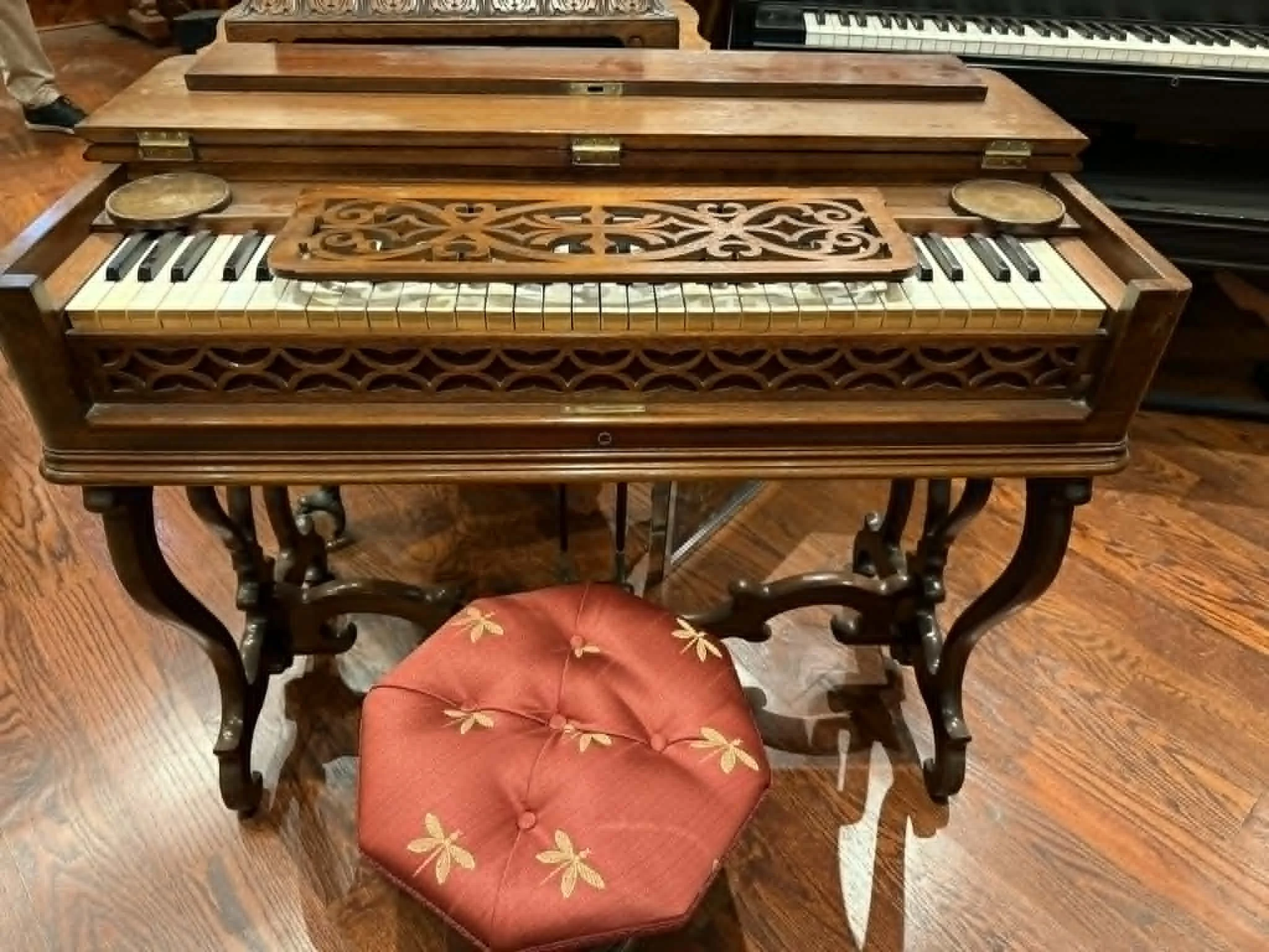 Antique wooden keyboard with intricate carvings, a sheet music stand, and a red upholstered stool with yellow floral embroidery.