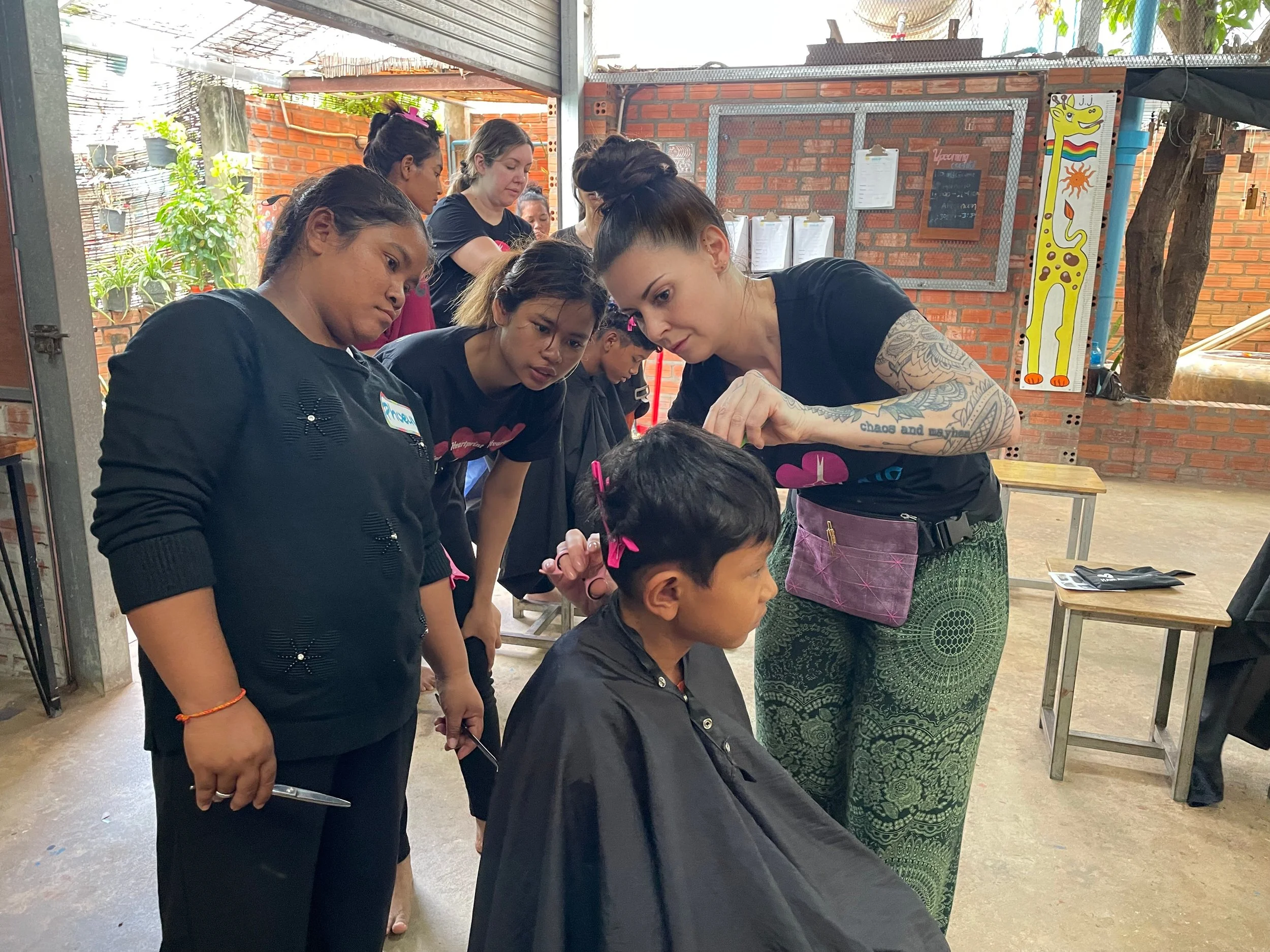 A woman with tattoos on her arm is giving a haircut to a young boy, while several women watch attentively in a salon or barbershop.