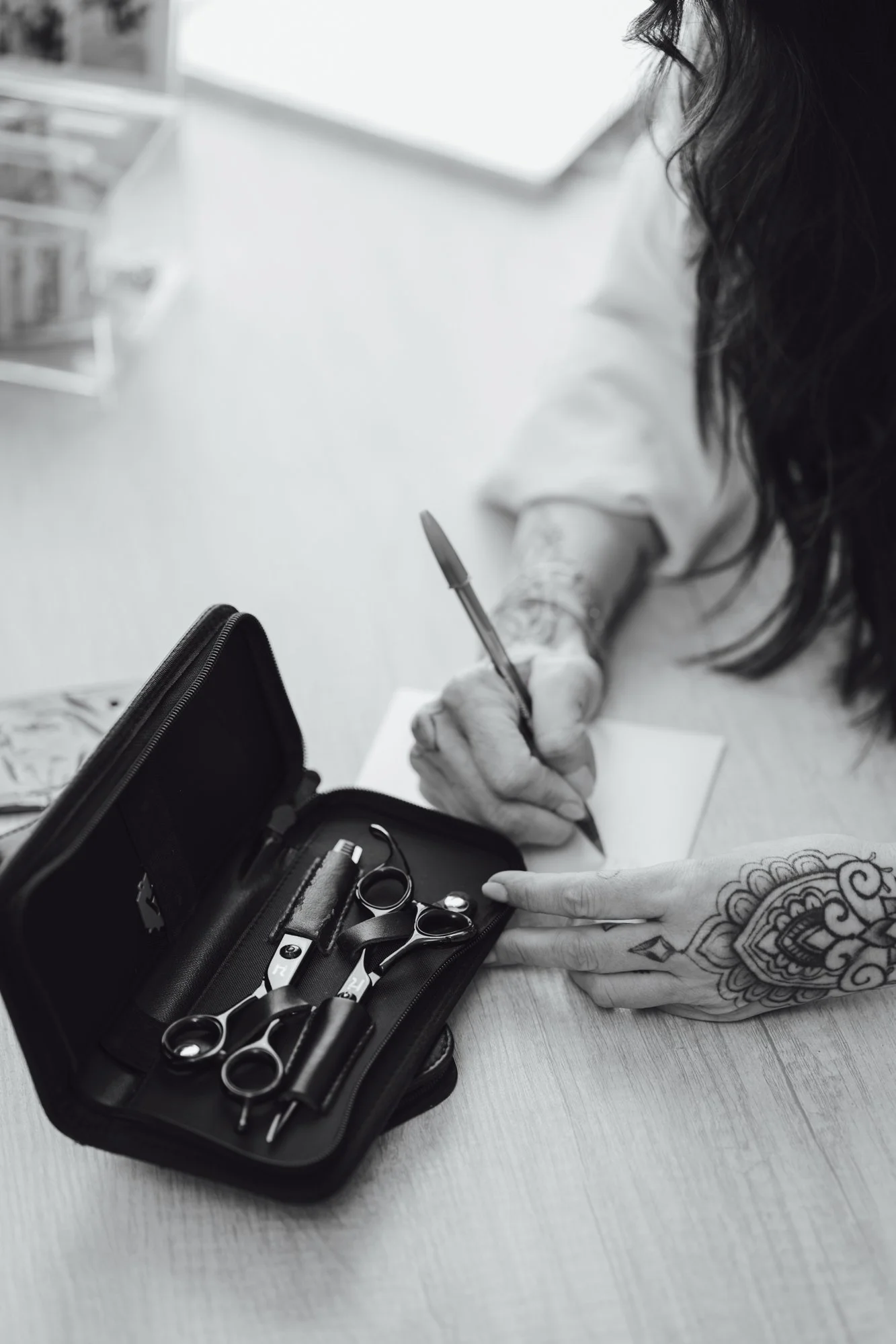 A person with tattoos on their arm writing with a pen on paper, with a manicure kit including scissors, nail clipper, and other tools on a black case on the table.