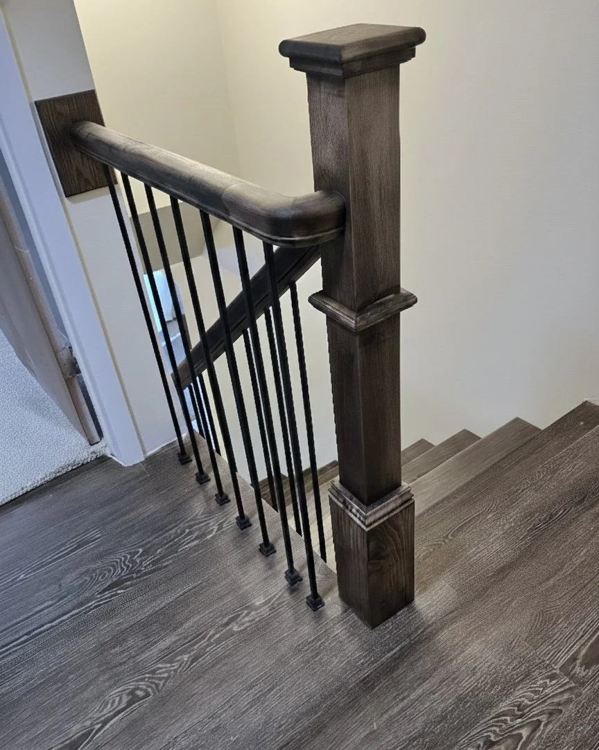 Wooden staircase railing with a thick, rounded handrail and metal balusters, adjacent to a wall and a door frame.