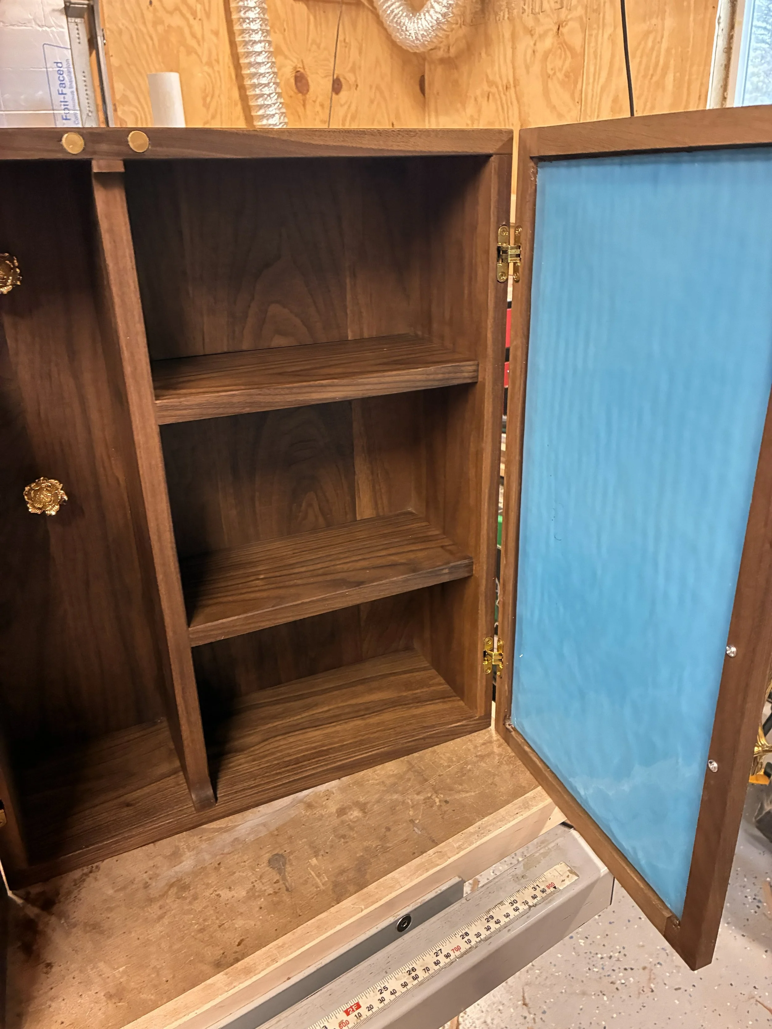 Empty wooden cabinet with blue backing, open door, in a workshop or garage.