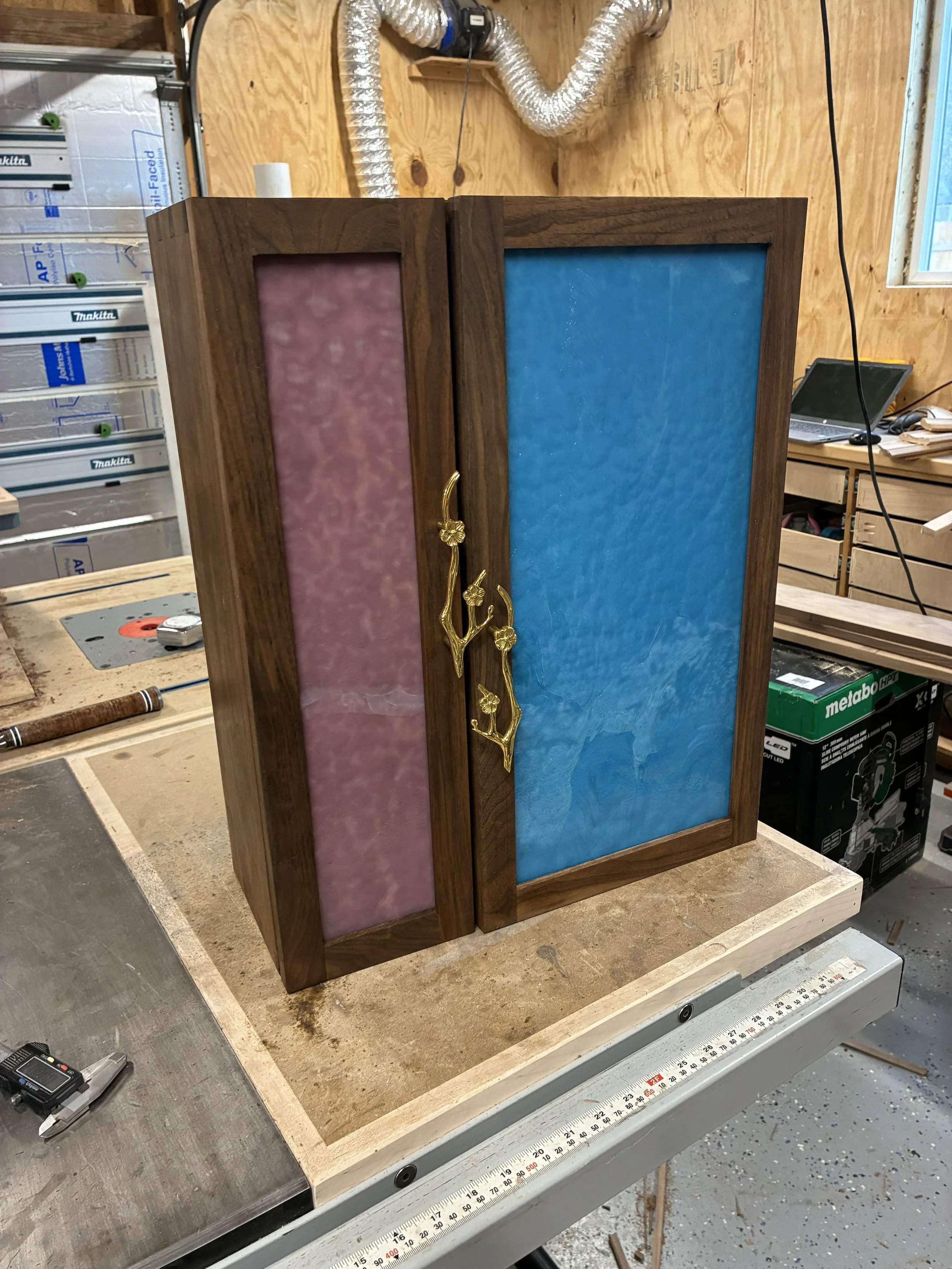 A wooden display cabinet with two colorful textured panels, one pink on the left and one blue on the right, with a gold decorative branch mounted on the left panel. The cabinet is situated in a workshop with tools and equipment in the background.