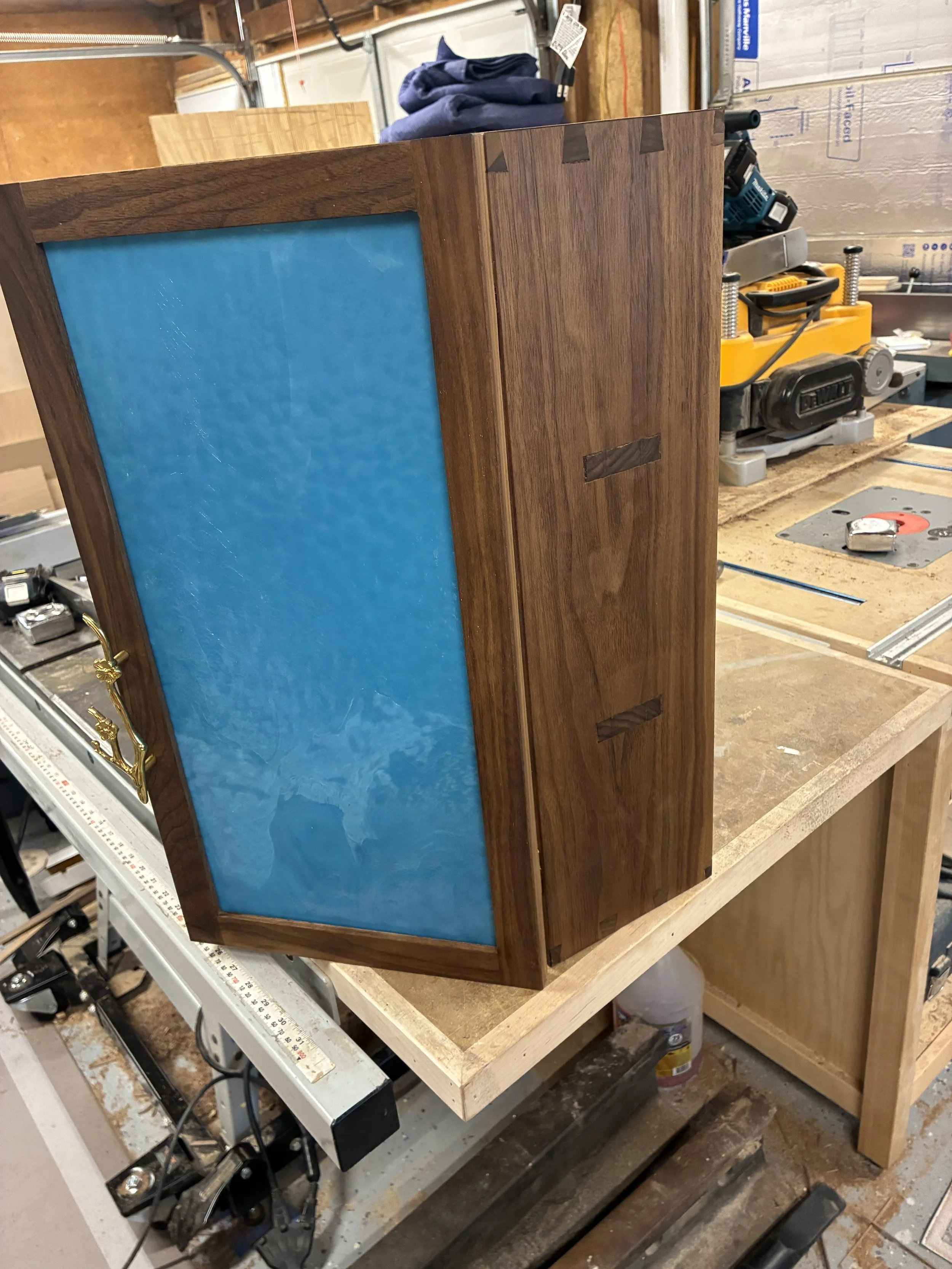 A small wooden cabinet with a blue panel on the front, situated on a workbench in a woodworking shop. The cabinet has a brass handle on the side, visible on the left side of the image.