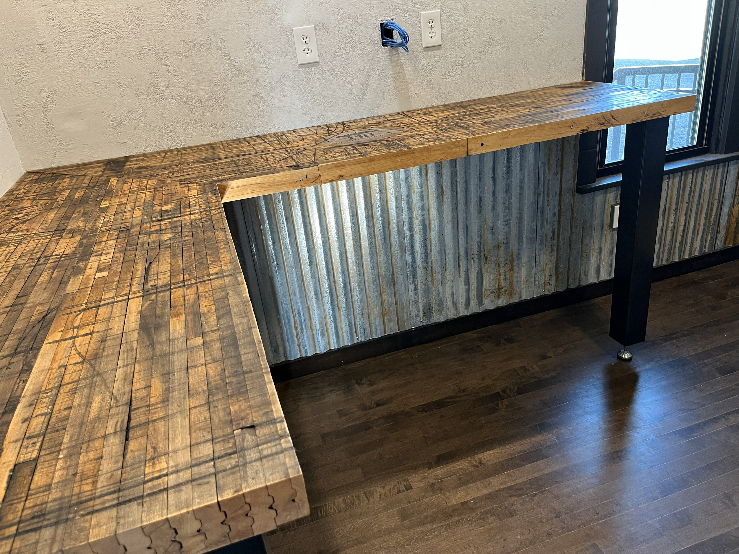 A rustic wooden bar counter with a metal corrugated panel beneath it, situated in front of a window with a balcony outside. The counter has a natural, rough finish with visible wood grain and some markings. There are three electrical outlets and a cu