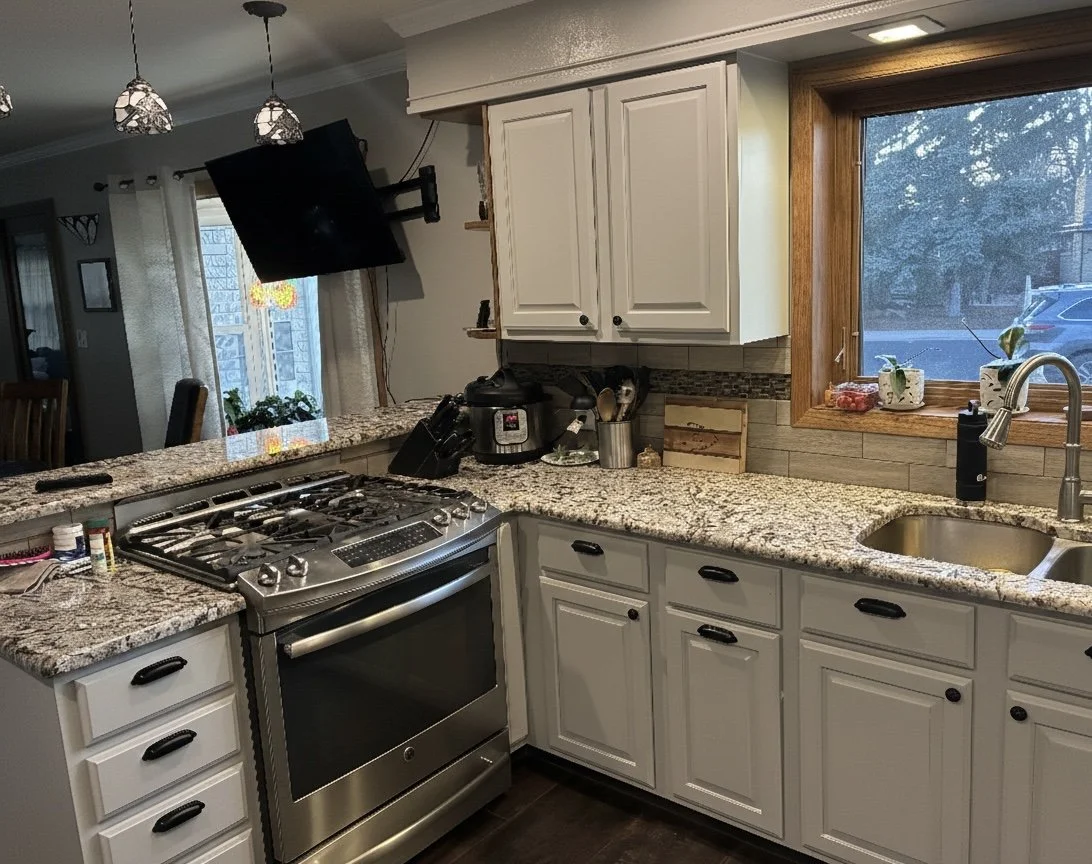 A kitchen with white cabinets, granite countertops, and a stainless steel stove. There is a window above the sink, a black flat-screen TV mounted on the wall, and various kitchen appliances and decor on the counter.