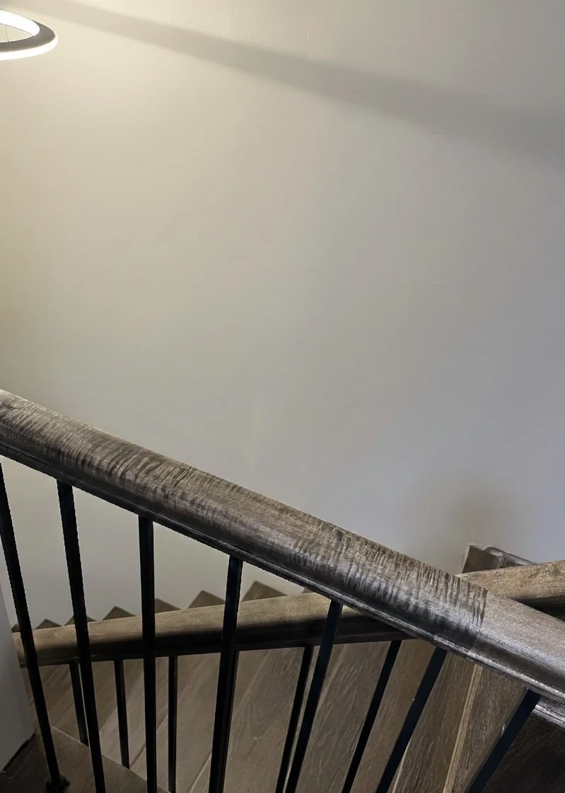 Photo of a staircase viewed from above, showing a wooden handrail and black metal balusters, with a plain white wall in the background.
