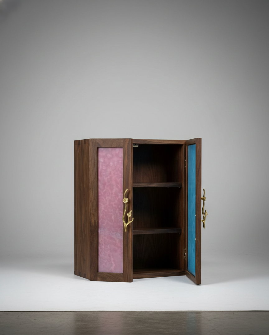 A small wooden cabinet with colorful panels on the doors, pink on the left and blue on the right, with gold-colored handles shaped like branches, placed on a white surface against a plain gray background.