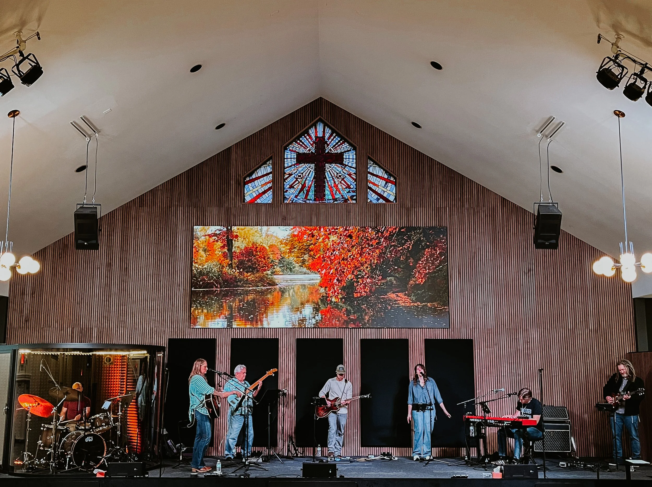 A band performs on stage inside a church with wooden vertical paneling, a large framed photo of fall foliage on the back wall, a stained glass window with a cross above it, and hanging lights.