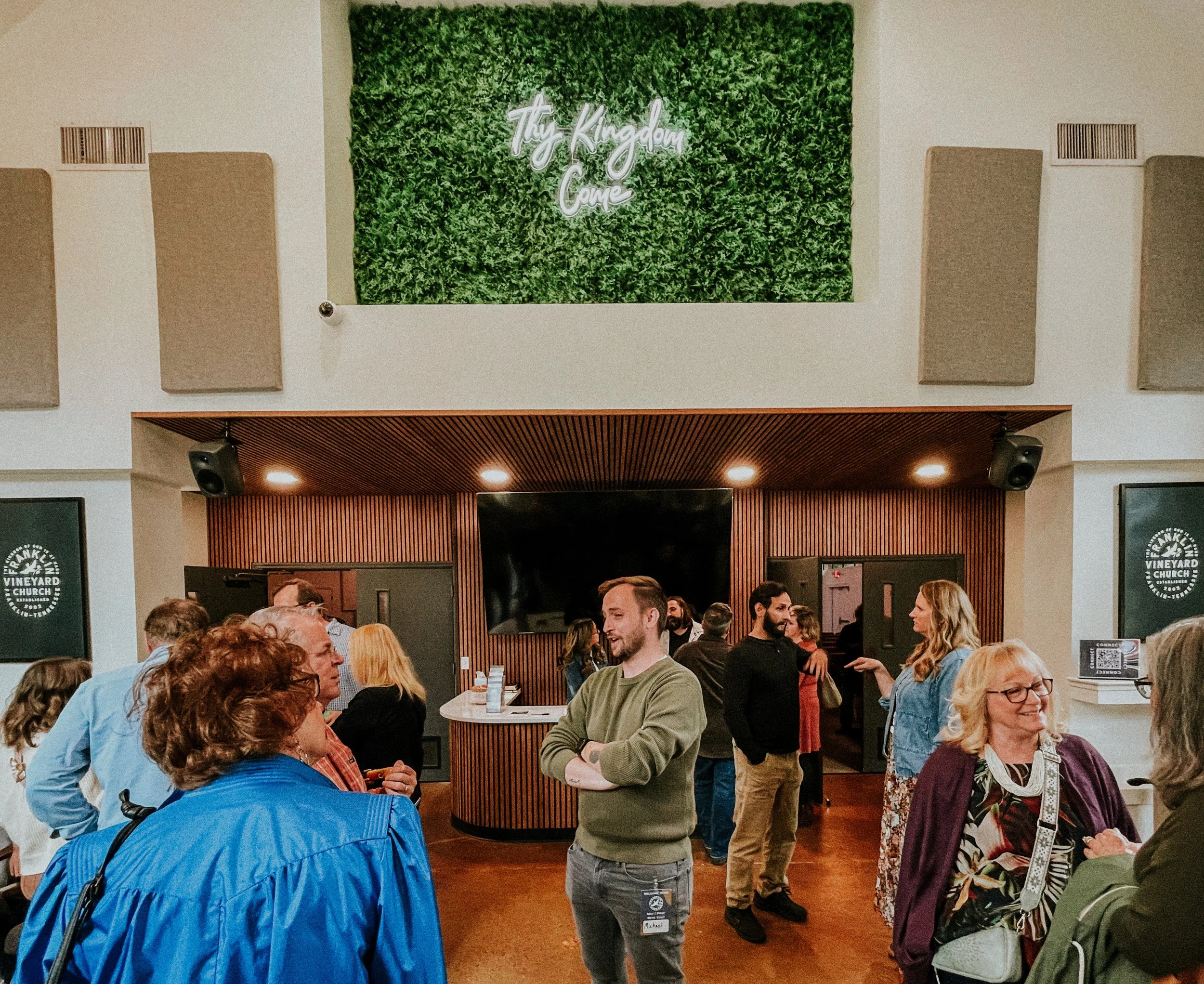People gathered inside a church with a sign that reads 'Tiny Kingdom Church' on a green wall.