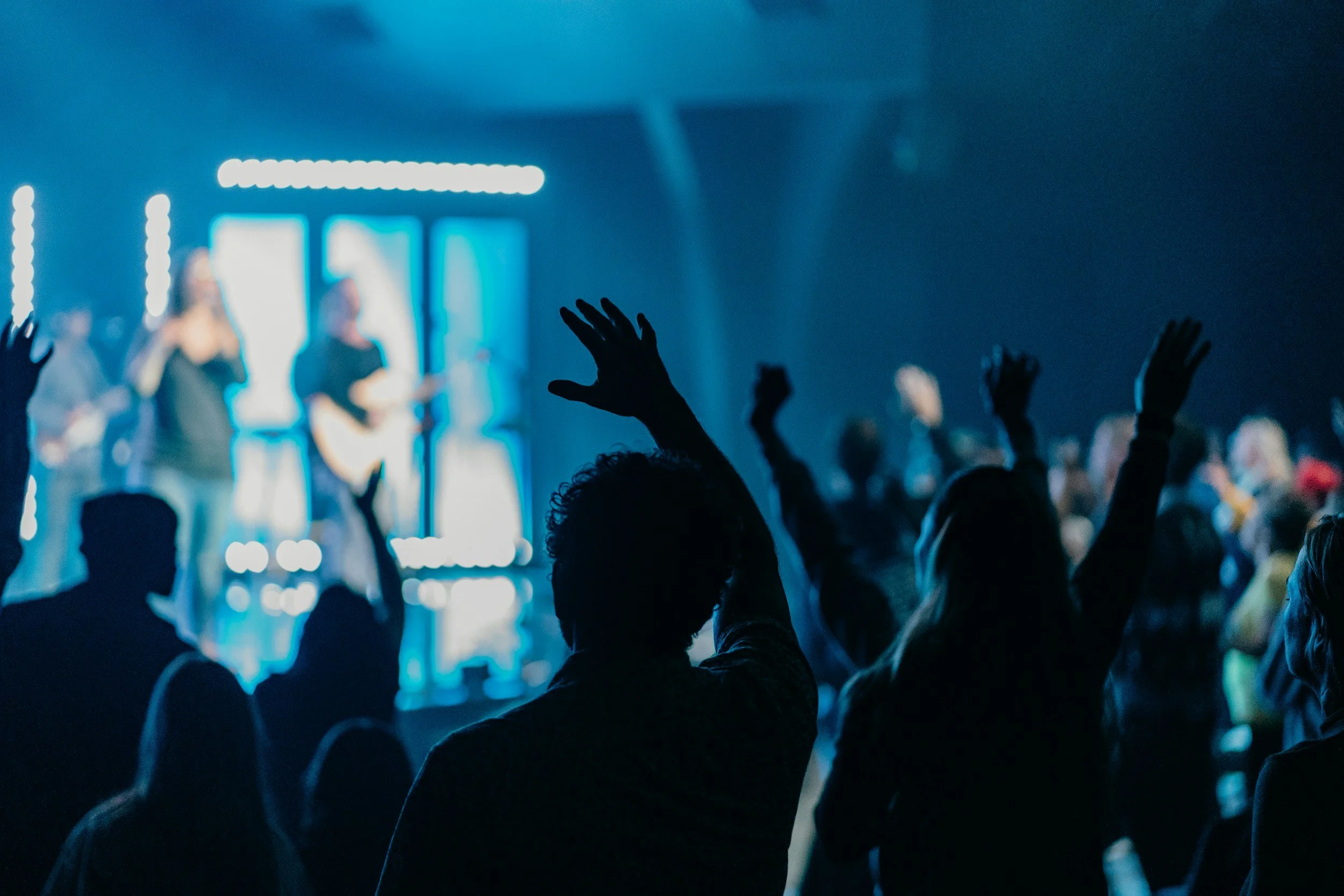 Silhouettes of people raising their hands in front of a stage with musicians performing under blue lighting.