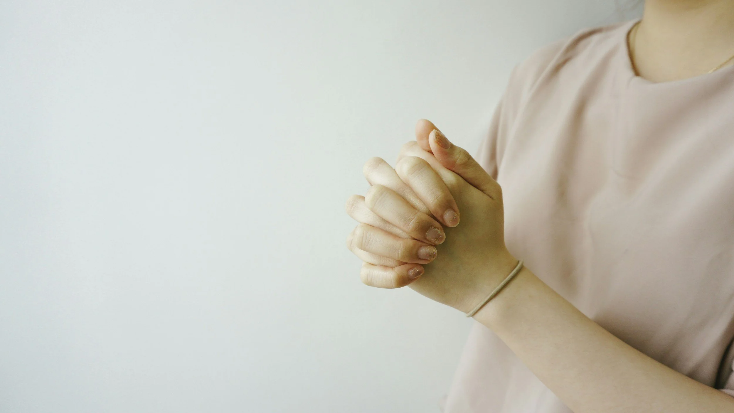 Person with light skin wearing a pink shirt and a bracelet, holding their hands together in a praying or meditative position against a plain background.