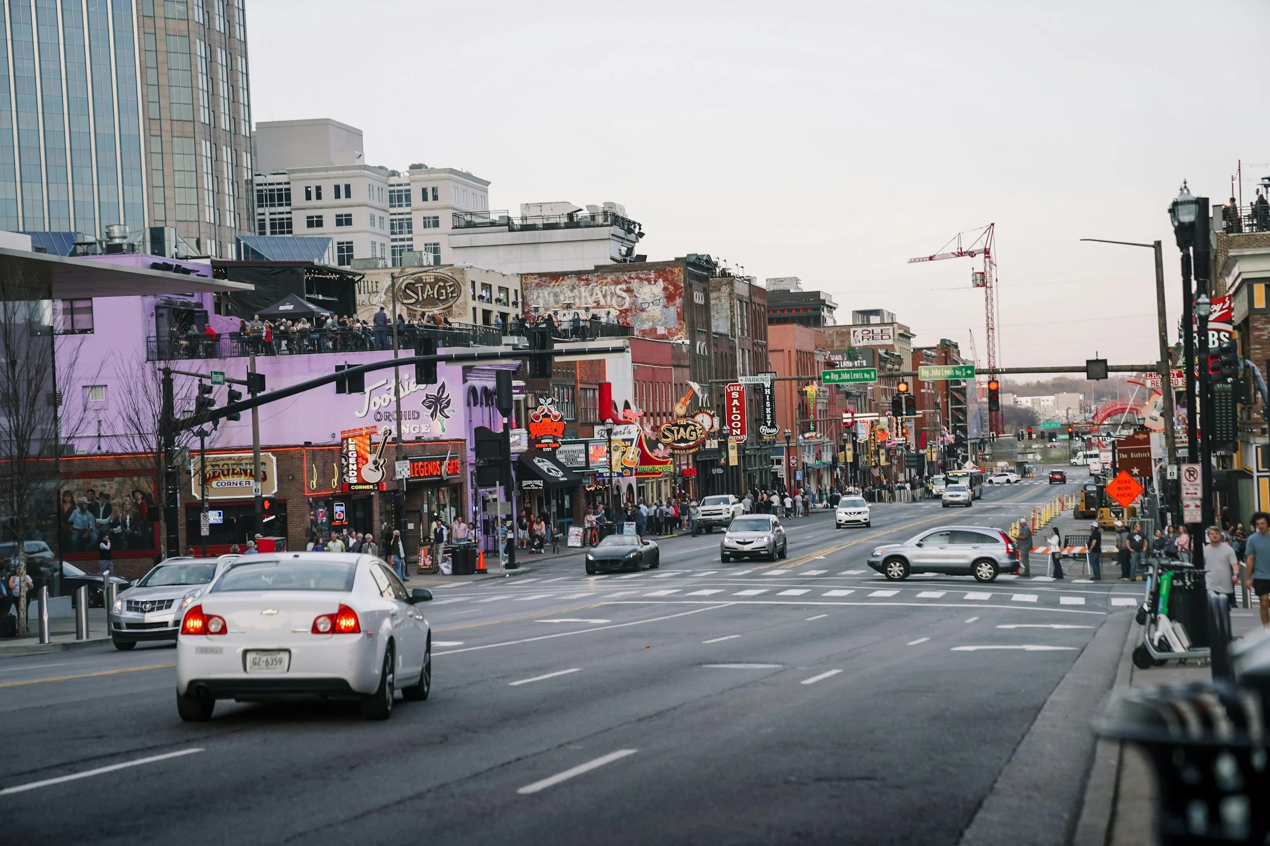 A city street in Nashville, Tennessee, with multiple shops, bars, and signs along the sidewalk. Cars are driving through the intersection, and pedestrians are walking on the crosswalk. Buildings and cranes are visible in the background.