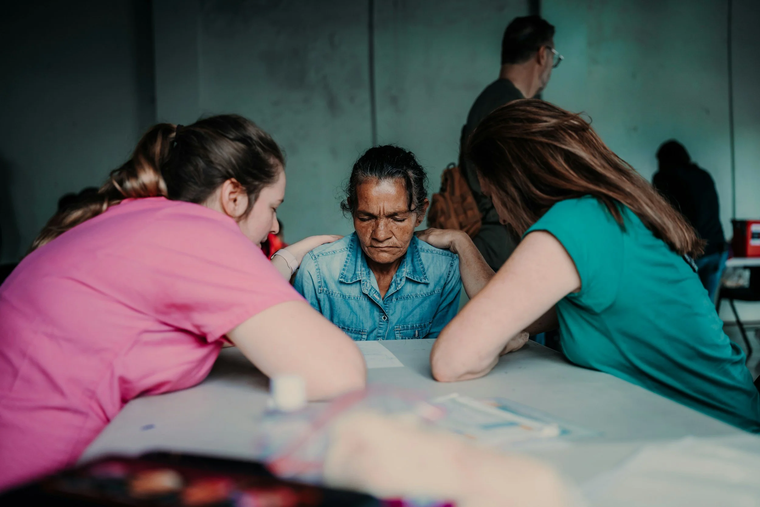 Two young women are offering comfort to a middle-aged woman who appears distressed, in a setting that resembles a support or counseling center.