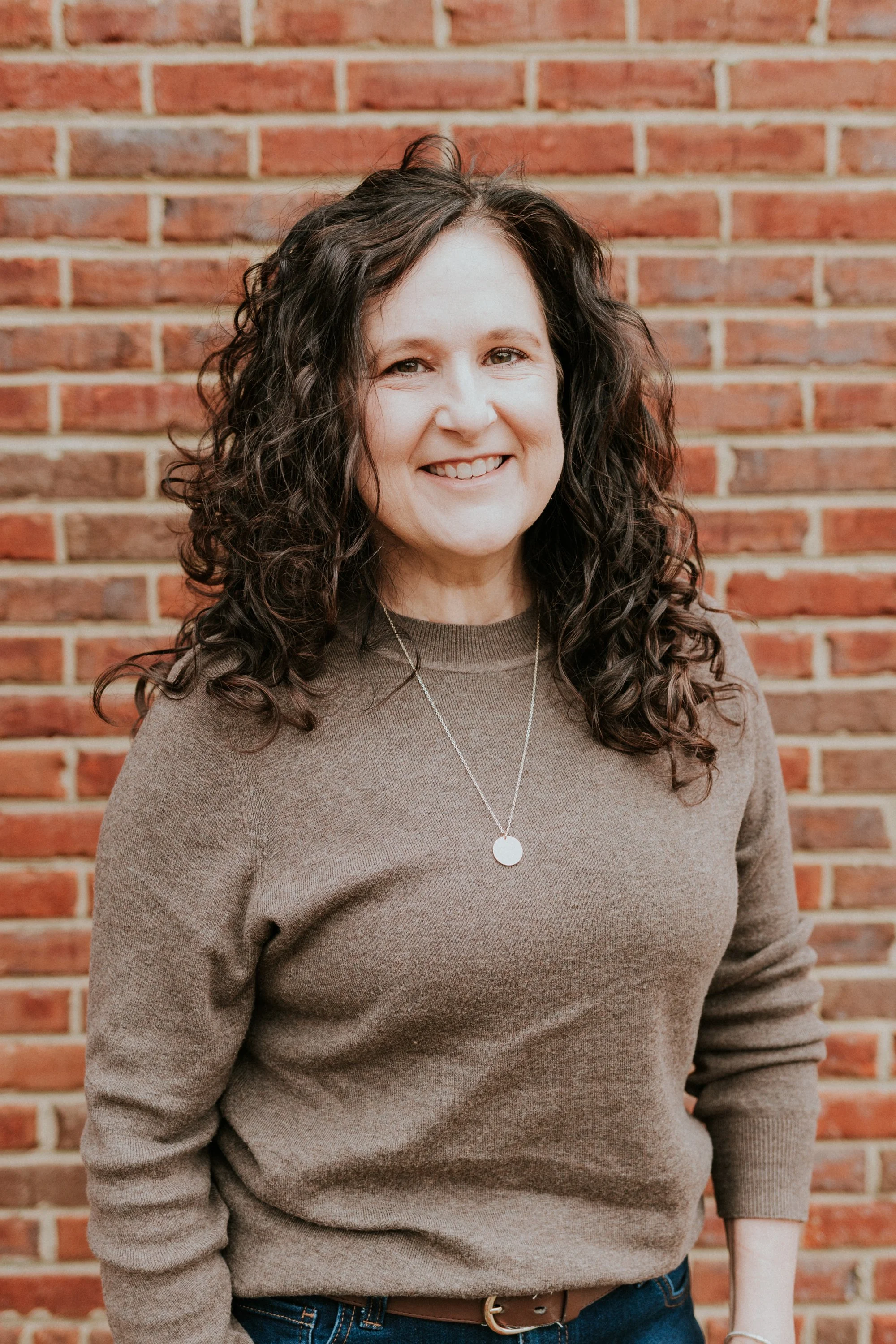 A woman with curly dark hair smiling in front of a brick wall, wearing a brown sweater and a silver necklace.