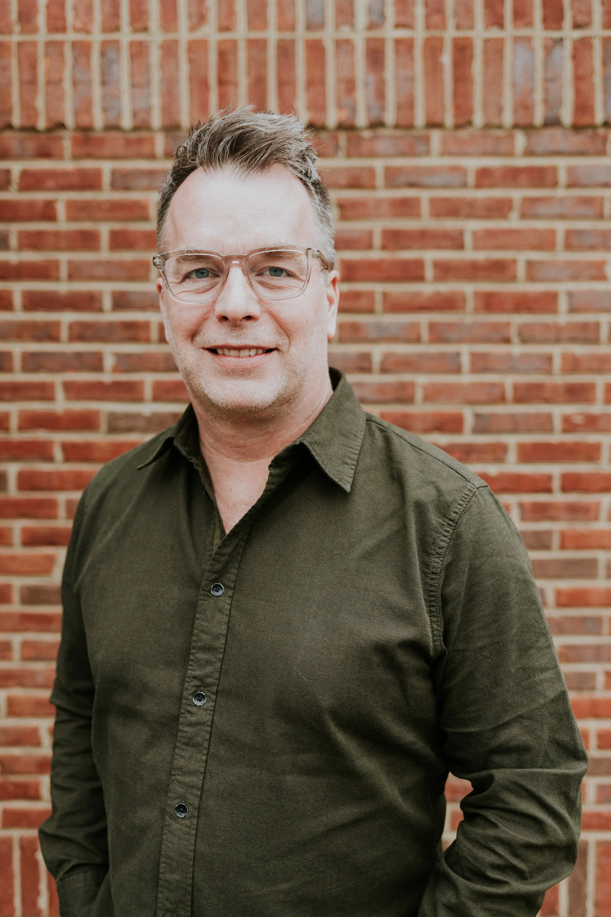 A man with glasses and short brown hair, wearing an olive green button-up shirt, standing outdoors in front of a red brick wall, smiling at the camera.