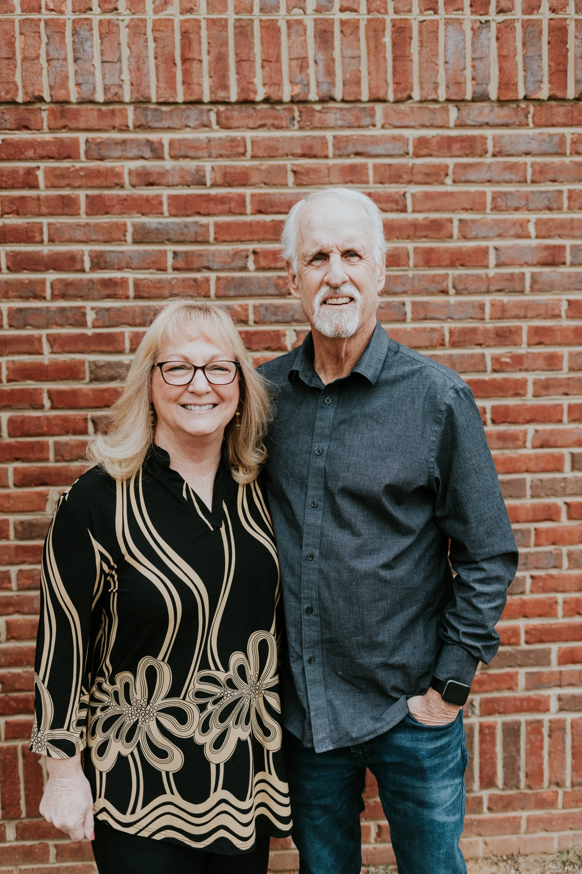 A smiling woman with blonde hair and glasses standing beside a serious-looking man with white hair and a beard, both standing in front of a red brick wall.