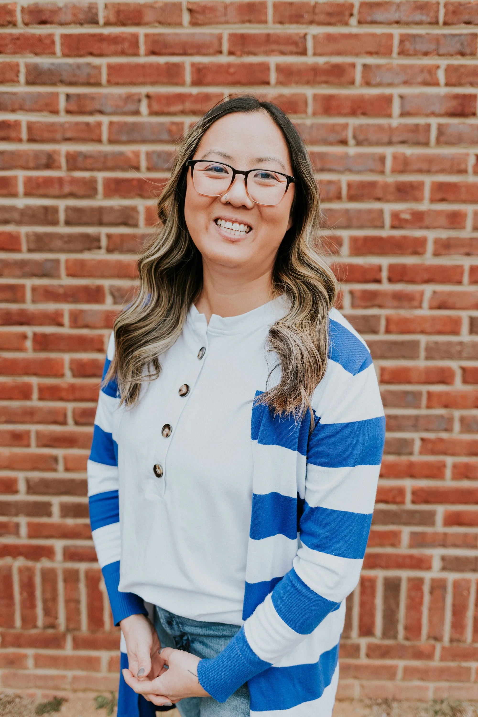 A smiling woman wearing glasses, a white shirt with black buttons, and a blue-and-white striped cardigan standing in front of a brick wall.