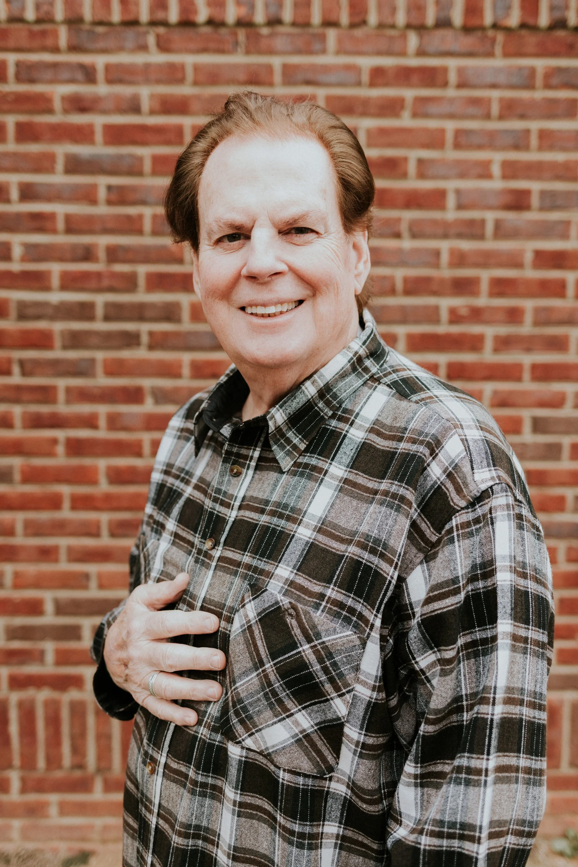 A man smiling, wearing a plaid shirt, standing in front of a brick wall, with his hand on the chest.