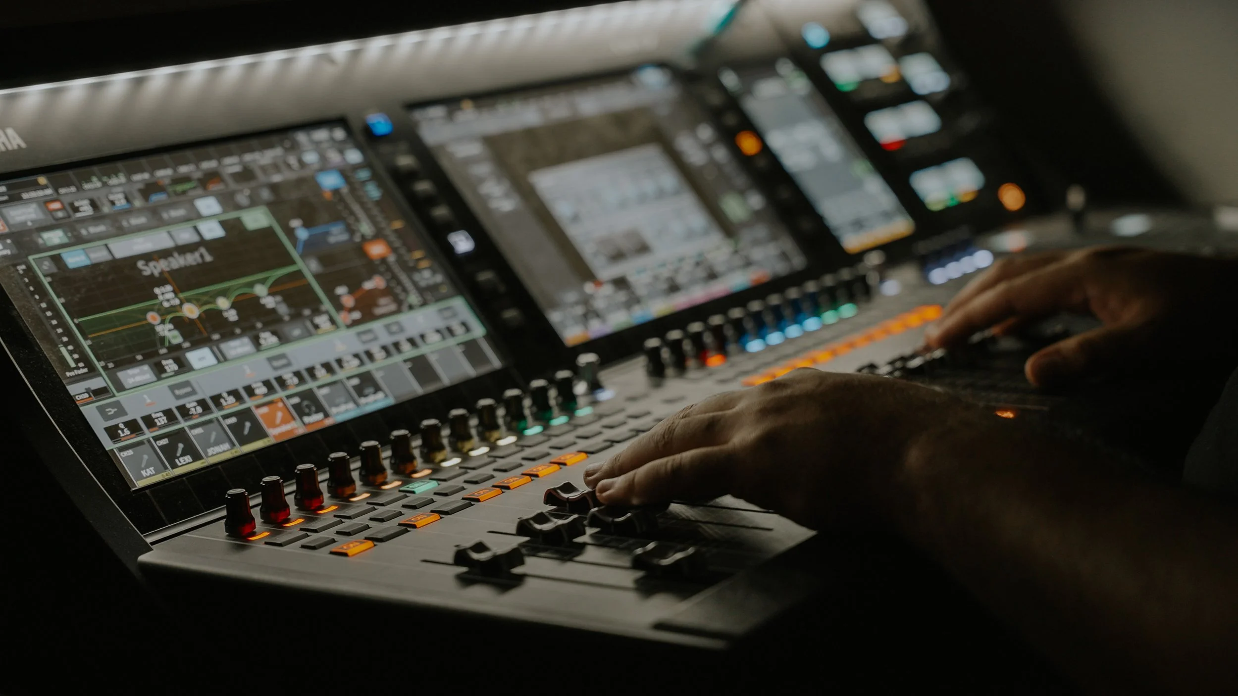Close-up of a person's hands operating a professional digital audio mixing console with multiple screens and colorful controls.